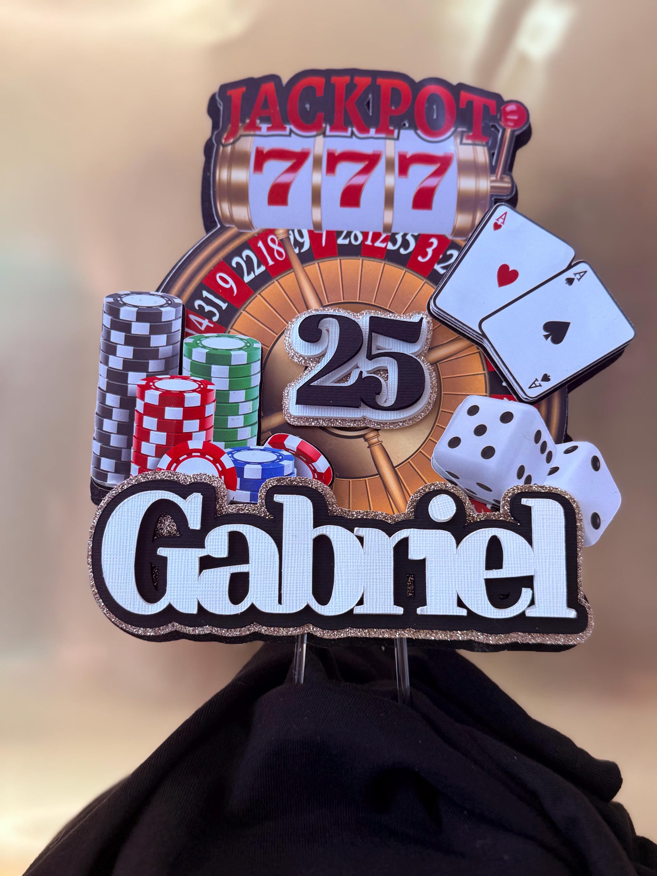 Personalized 3D cake topper with gambling-themed design and name 'Gabriel' on a blurred background