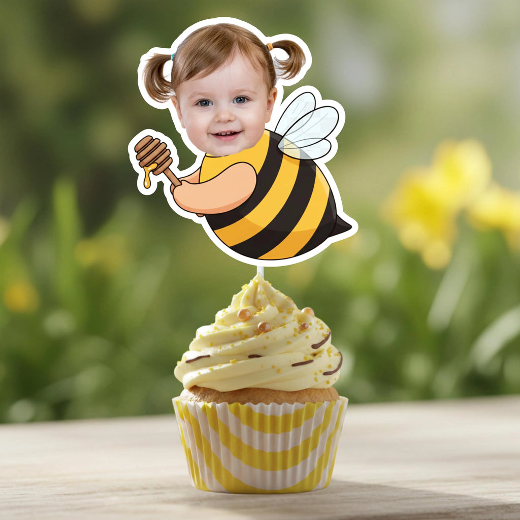 Cupcake with a bee-themed topper featuring a child's face, set against a natural background.