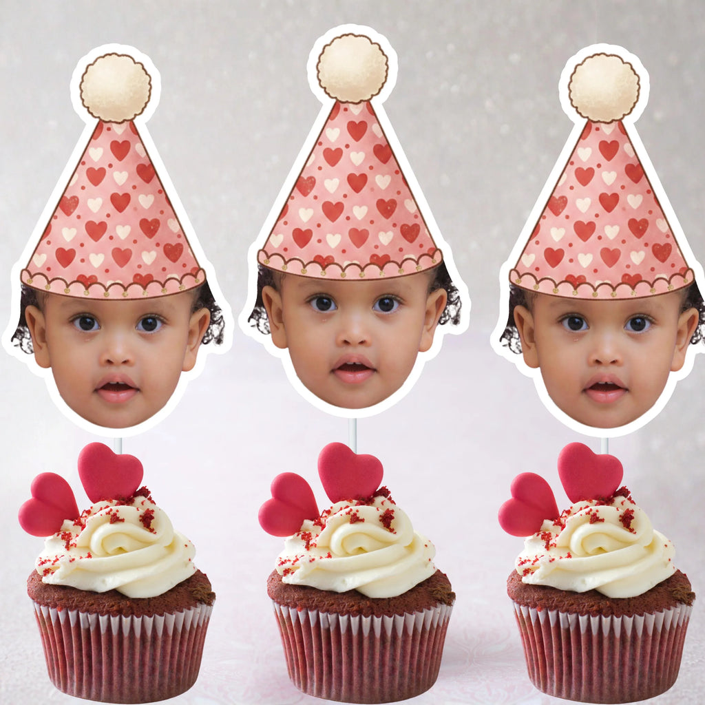 Three cupcakes with red and white frosting and pink heart decorations, each topped with a paper party hat featuring hearts and a child's face.