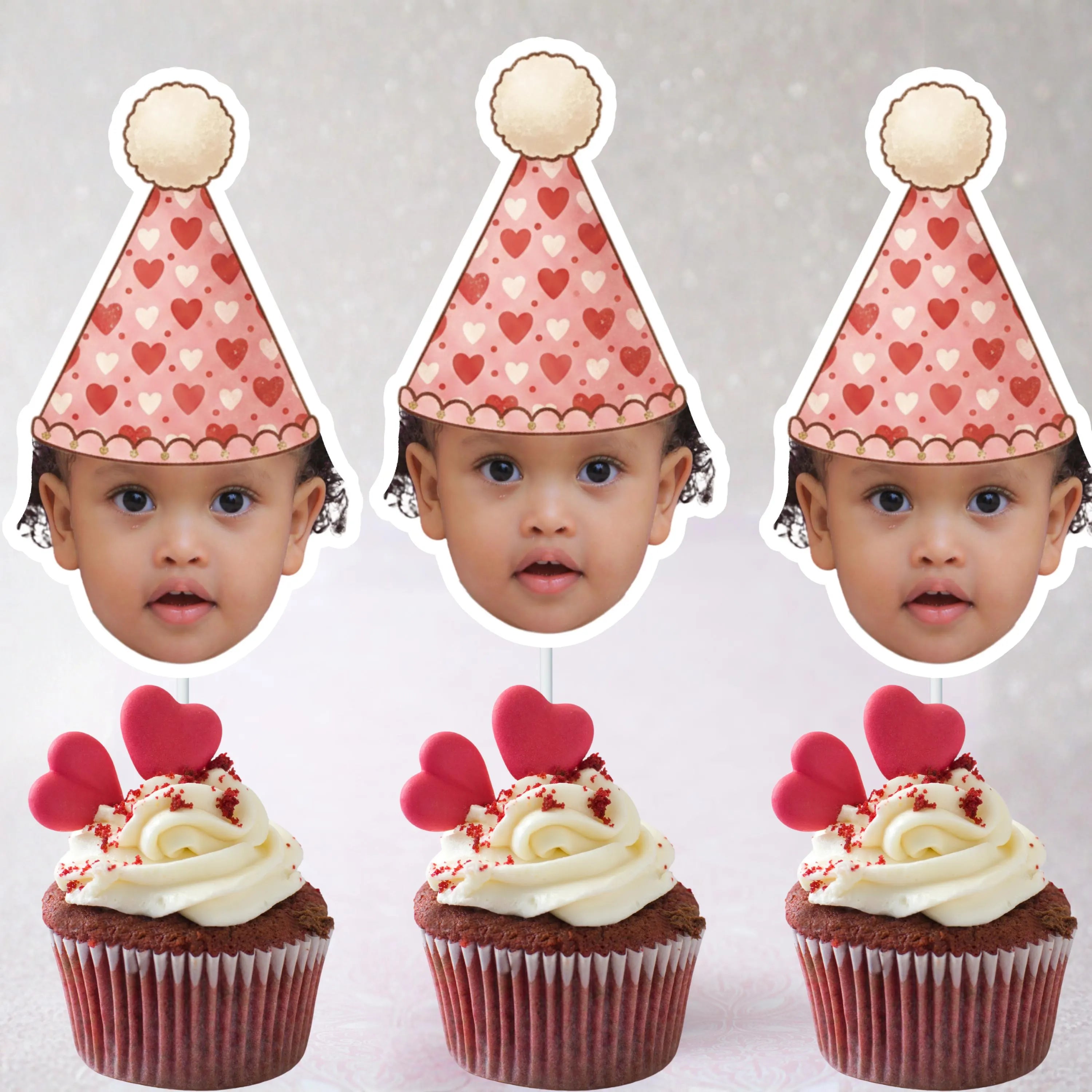 Three cupcakes with red and white frosting and pink heart decorations, each topped with a paper party hat featuring hearts and a child's face.