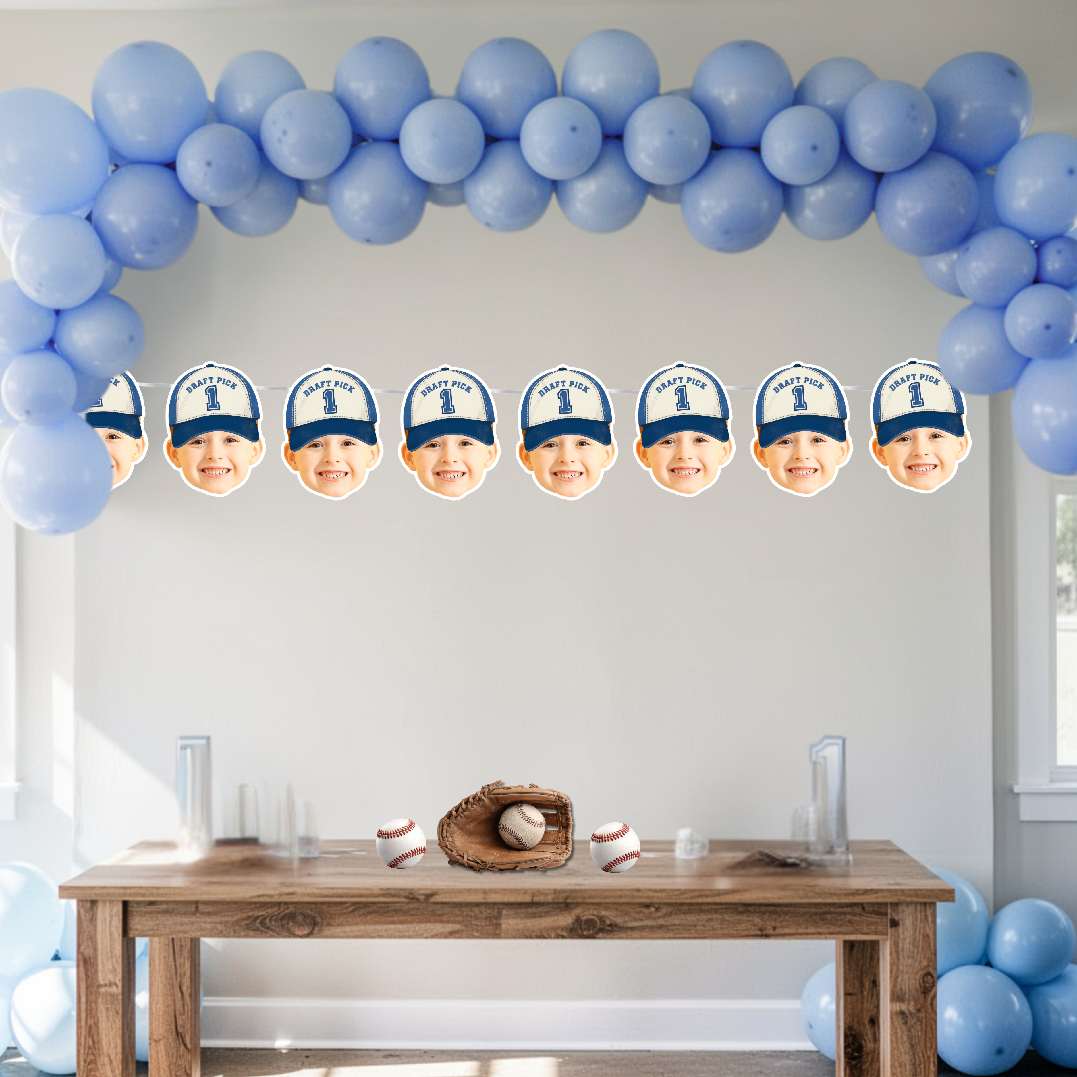 Personalized baseball first birthday banner with child’s photo wearing Draft Pick 1 cap, styled under blue balloon arch above dessert table with baseball glove and balls for Rookie of the Year sports themed party decorations.