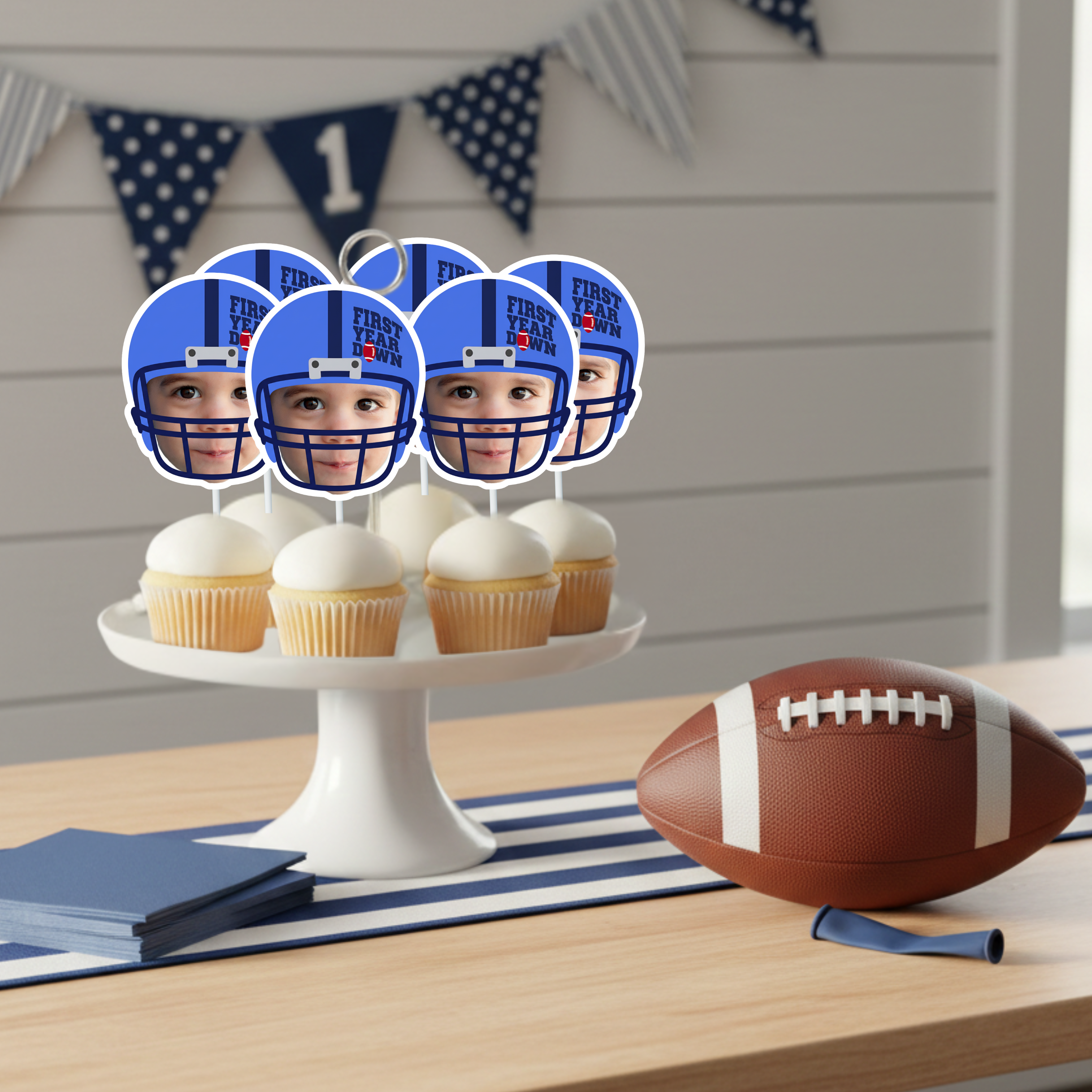 Football themed first birthday cupcake display featuring blue helmet personalized photo toppers arranged on white cake stand with football prop and navy banner background. Sports baby birthday party setup styled for rookie of the year game day celebration.