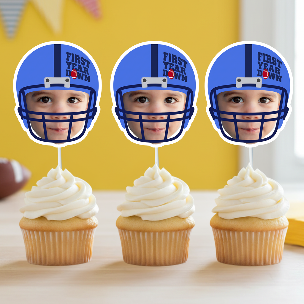 Three blue football helmet First Year Down personalized cupcake toppers featuring baby photo design displayed on vanilla cupcakes with smooth white frosting. Blue team color sports themed first birthday decorations styled for game day baby celebration.