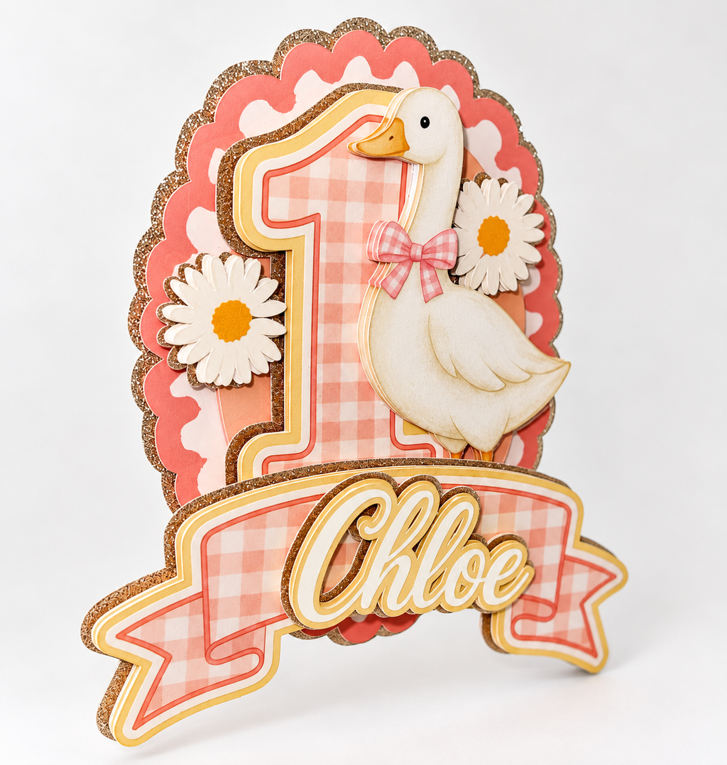 Side view of personalized Chloe first birthday goose cake topper showing layered laminated cardstock, pink gingham banner, white goose, daisies, and champagne gold glitter detailing.