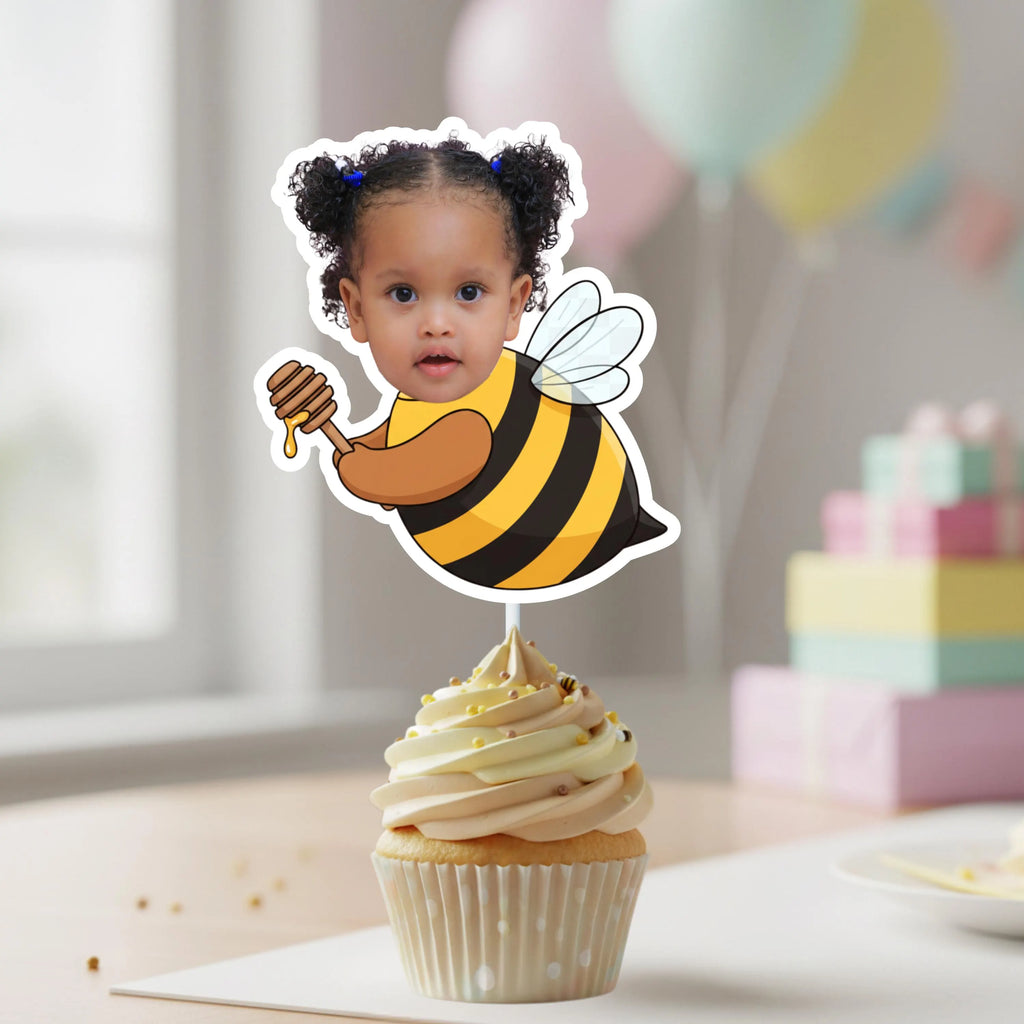Cupcake with a bee-themed topper featuring a child's face, with balloons and a cake in the background.