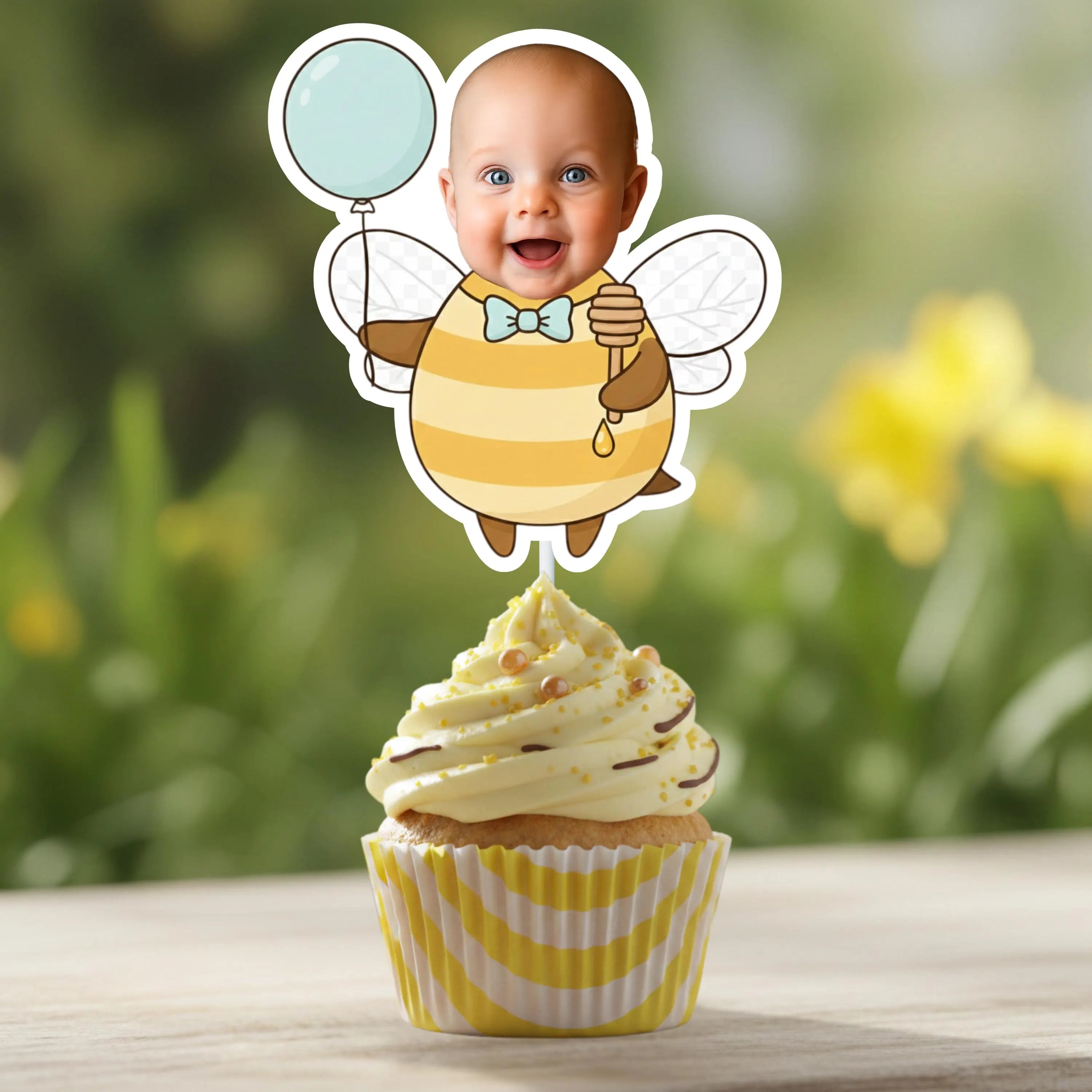 Cupcake with a bee-themed topper featuring a baby's face on a blurred natural background