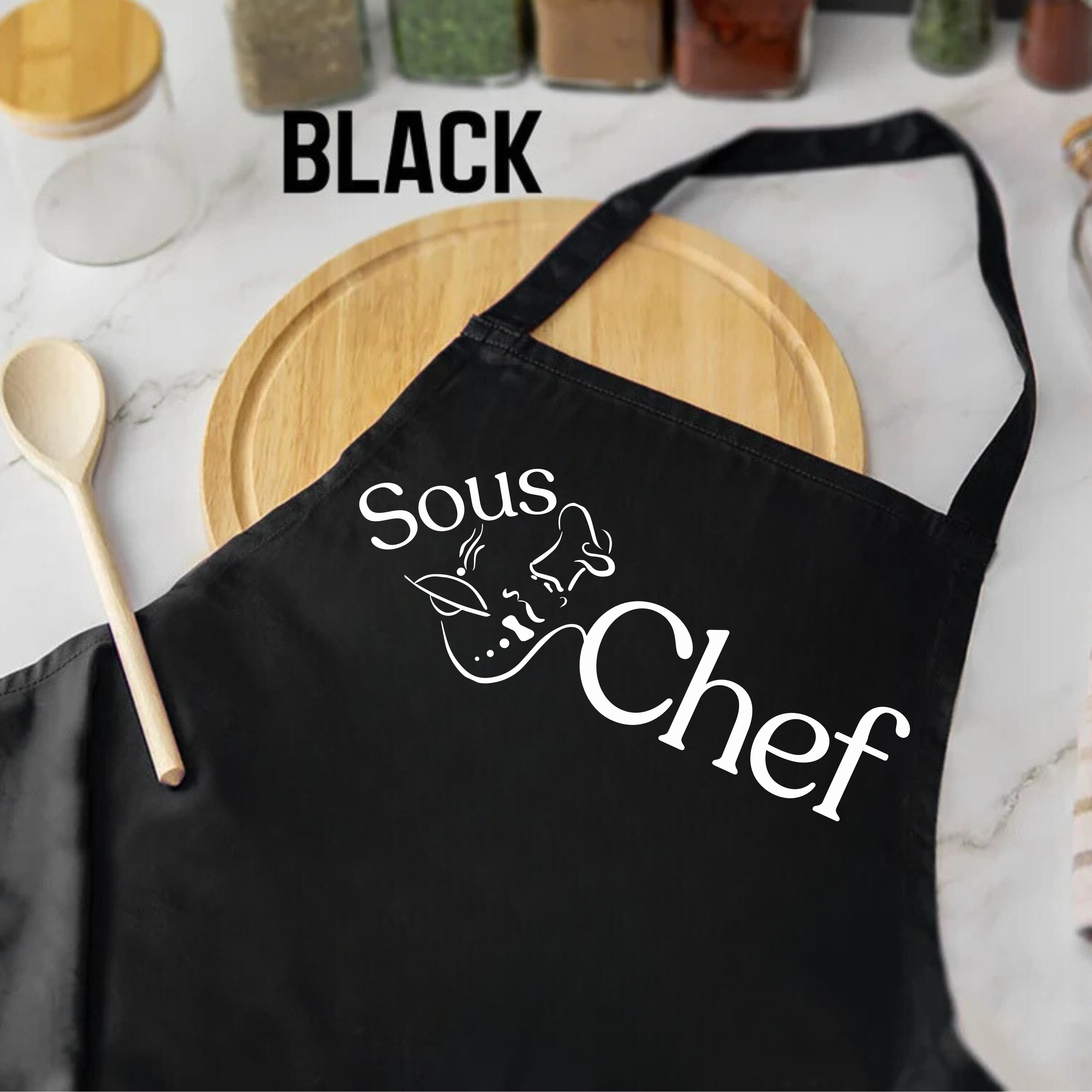 Black apron with 'Sous Chef' text on a kitchen counter with wooden cutting board and spoon.