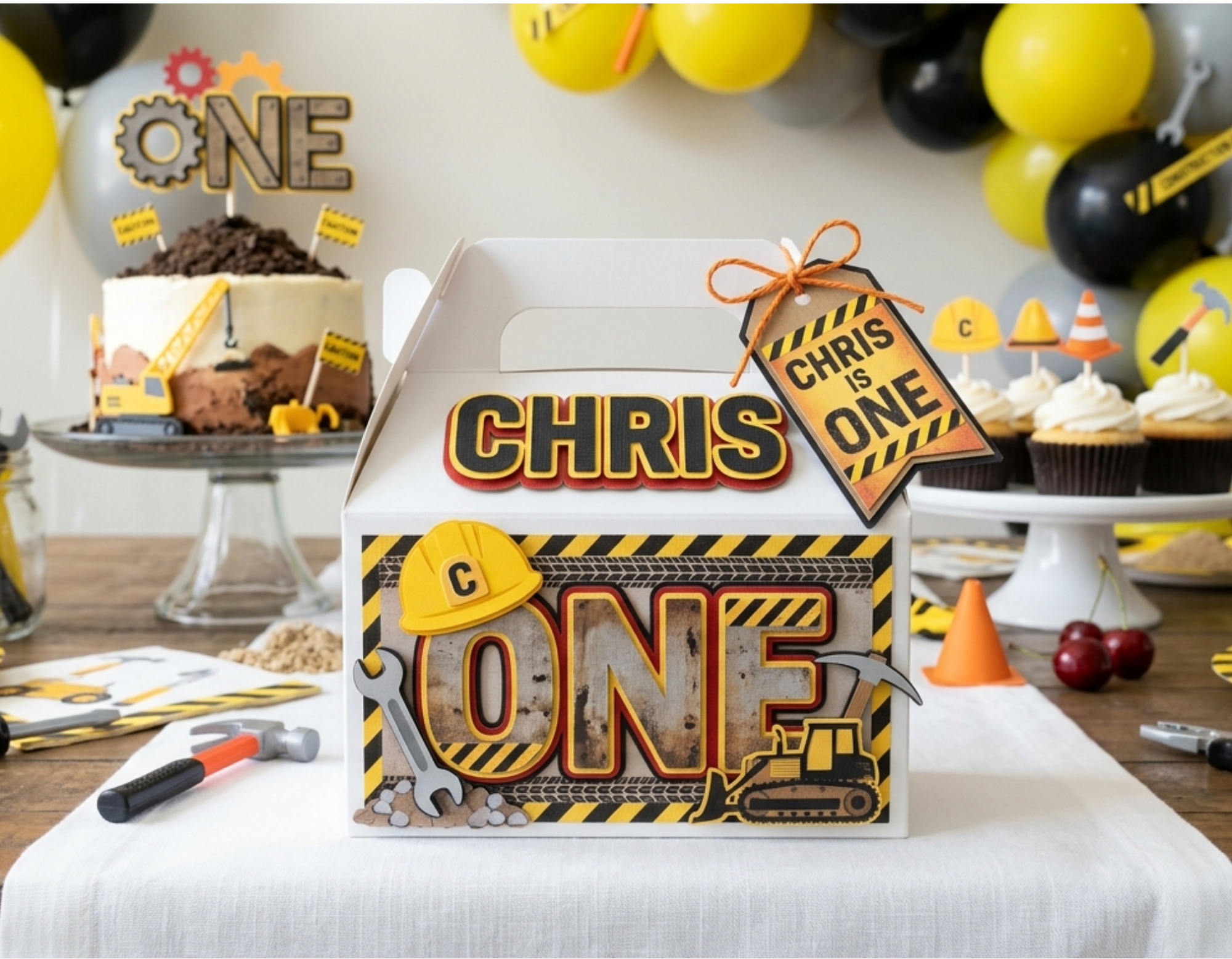 Construction themed birthday party setup featuring custom handmade party favor gable box, layered cardstock construction decorations, birthday cake, cupcakes, balloons, and coordinated first birthday party decor.