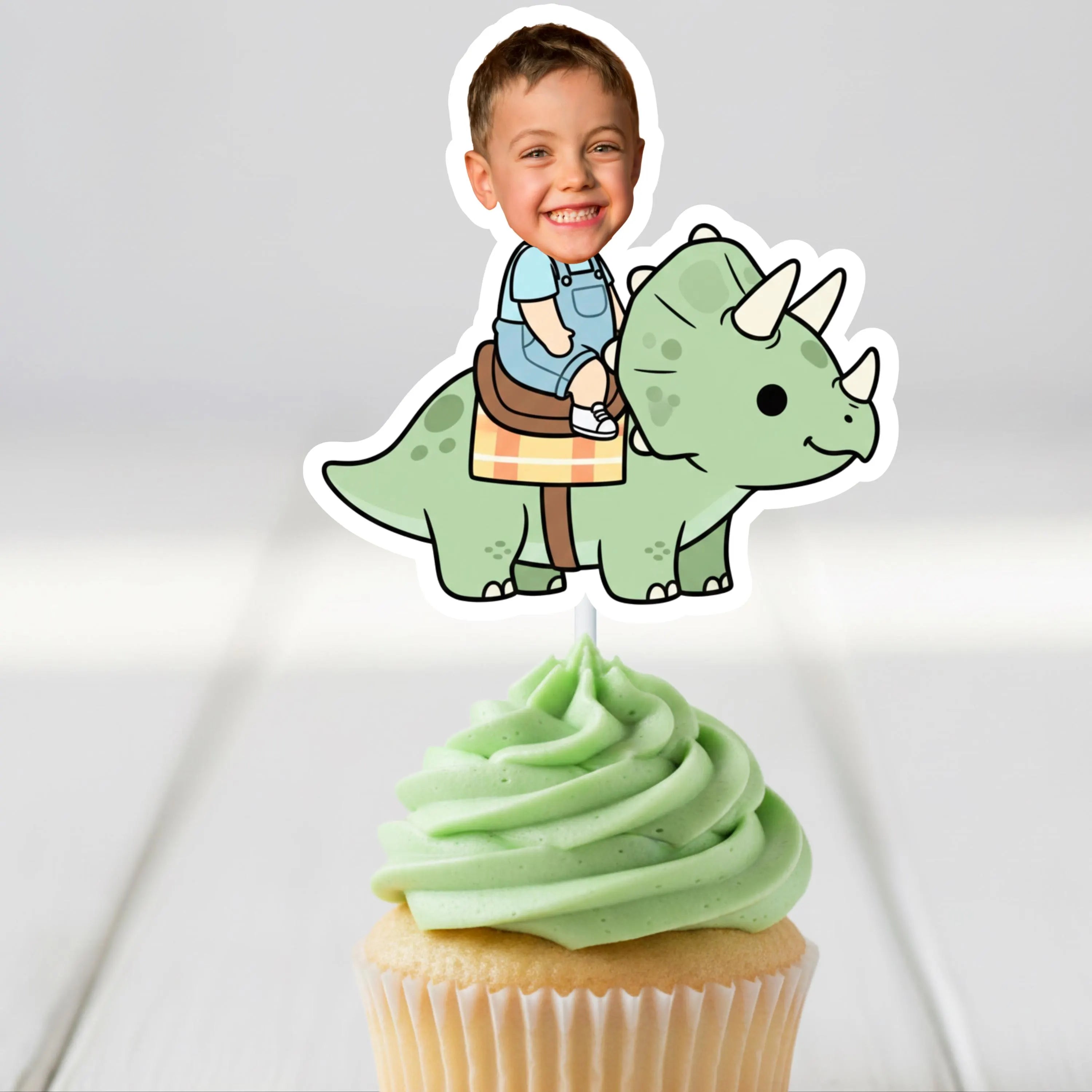 Cupcake with green frosting and a dinosaur-shaped cupcake topper with a child's face on a gray background