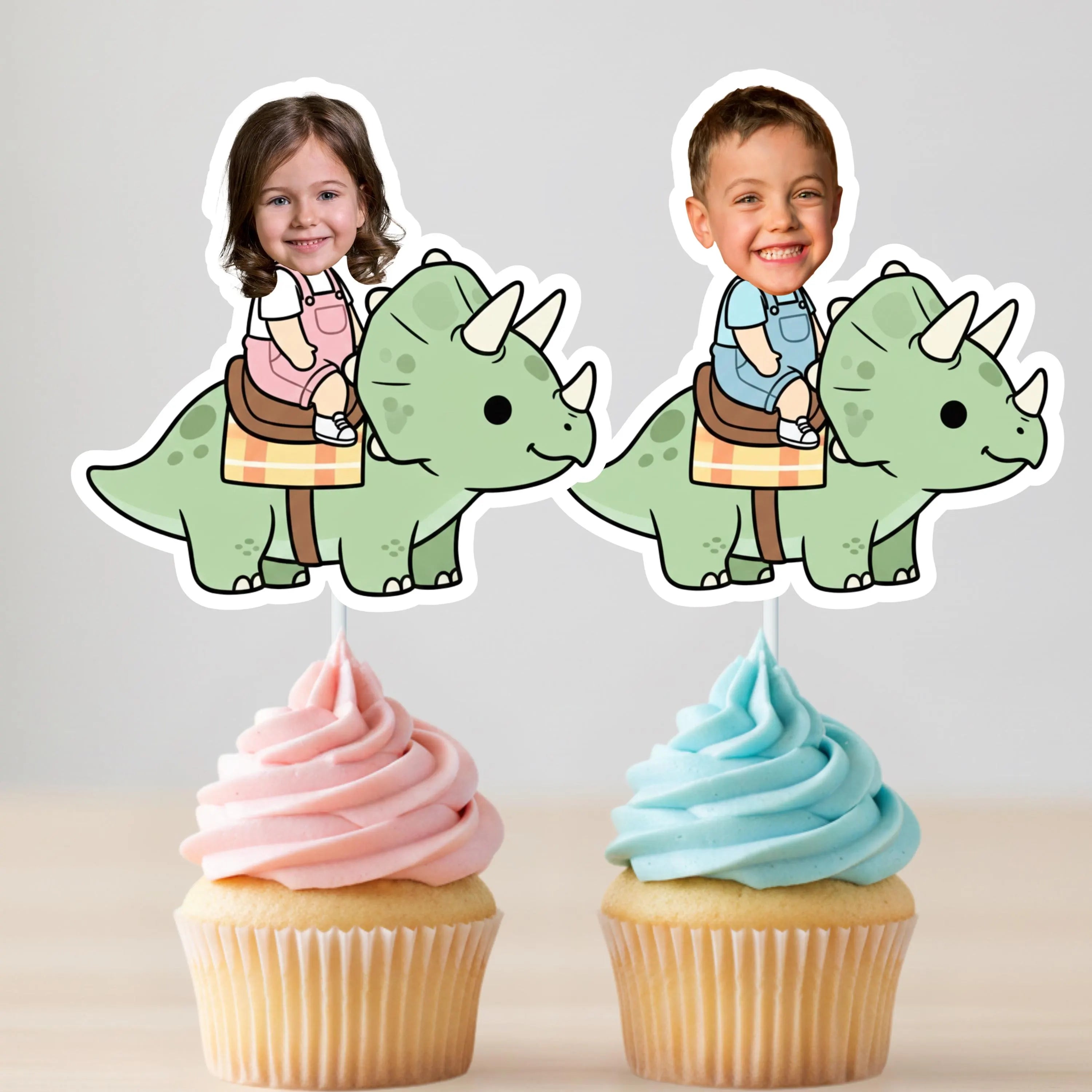 Two cupcakes with pink and blue frosting, each topped with a dinosaur cupcake topper featuring a child riding a dinosaur.