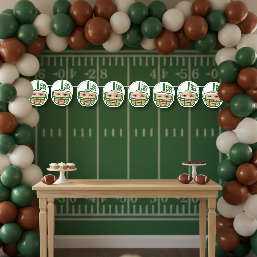 First Year Down football photo banner displayed against a football field backdrop with yard lines and balloon arch. Custom baby face helmet banner designed for a football themed first birthday party dessert table and game day celebration.