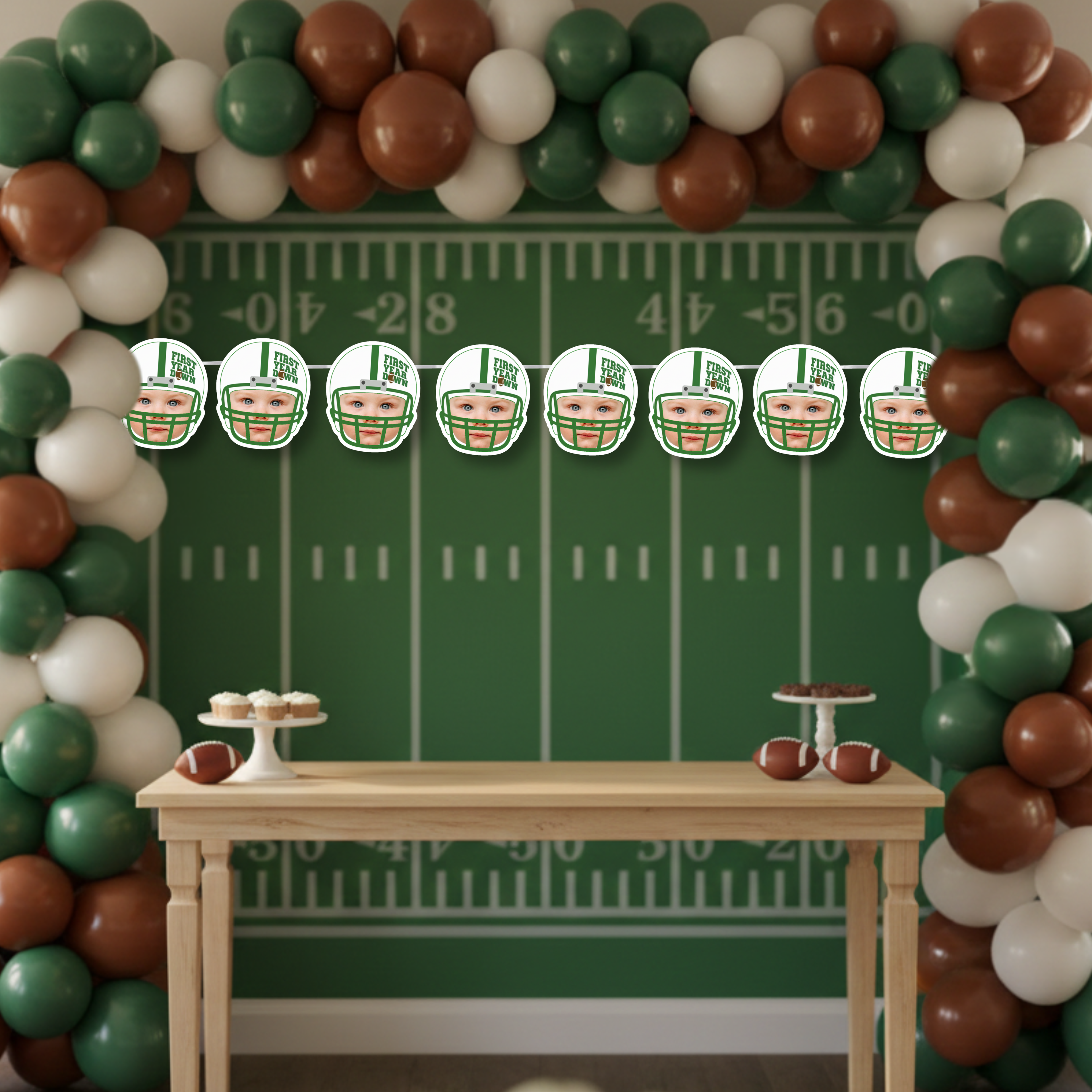 First Year Down football photo banner displayed against a football field backdrop with yard lines and balloon arch. Custom baby face helmet banner designed for a football themed first birthday party dessert table and game day celebration.
