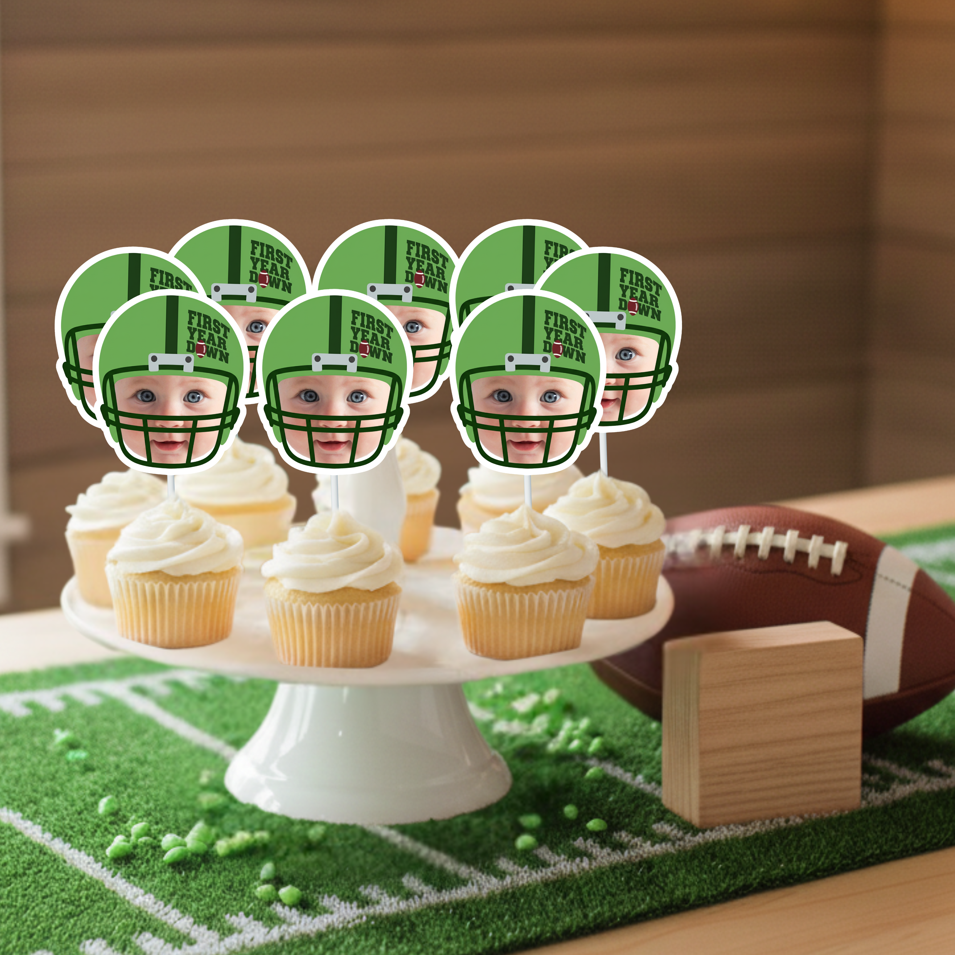 Football first birthday smash cake table styled with green turf field mat, football decoration, and personalized First Year Down cupcake toppers featuring baby photo design displayed on vanilla cupcakes. Sports themed one year old birthday party centerpiece idea.