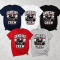 Five short-sleeve graphic T-shirts arranged on a light gray textured background, shown in navy, white, black, red, and heather gray. Each shirt features a football helmet and football graphic with the text 'GAME DAY CREW' and '2025'.