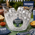 A clear plastic ice bucket filled with silver aluminum beverage cans, placed on a green football-field-themed table with snacks and football memorabilia around it. The center can features a personalized round sticker reading 'THE JOHNSON'S WATCH PARTY' with a football helmet graphic.