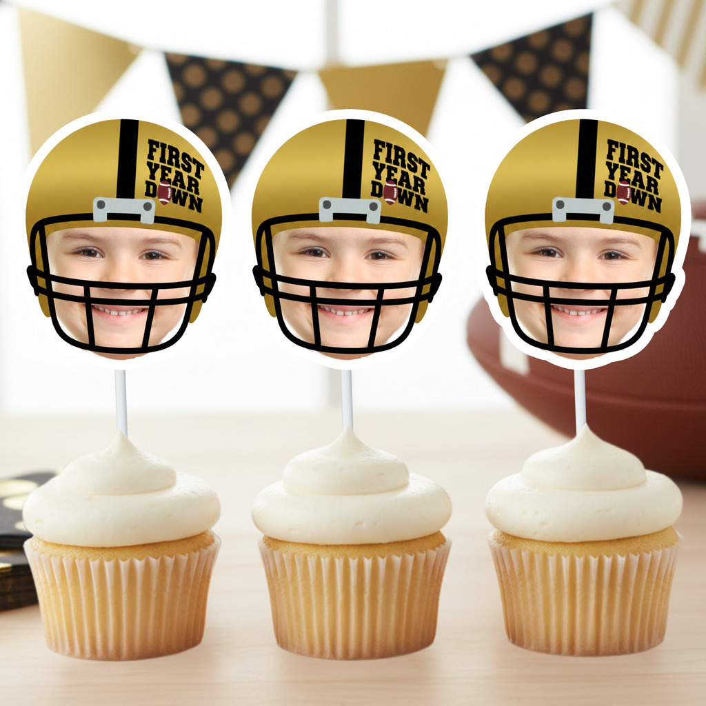 Three gold football helmet First Year Down custom photo cupcake toppers displayed on frosted cupcakes for football themed first birthday celebration. Gold and black sports birthday decor styled with football and banner backdrop for rookie of the year baby party.