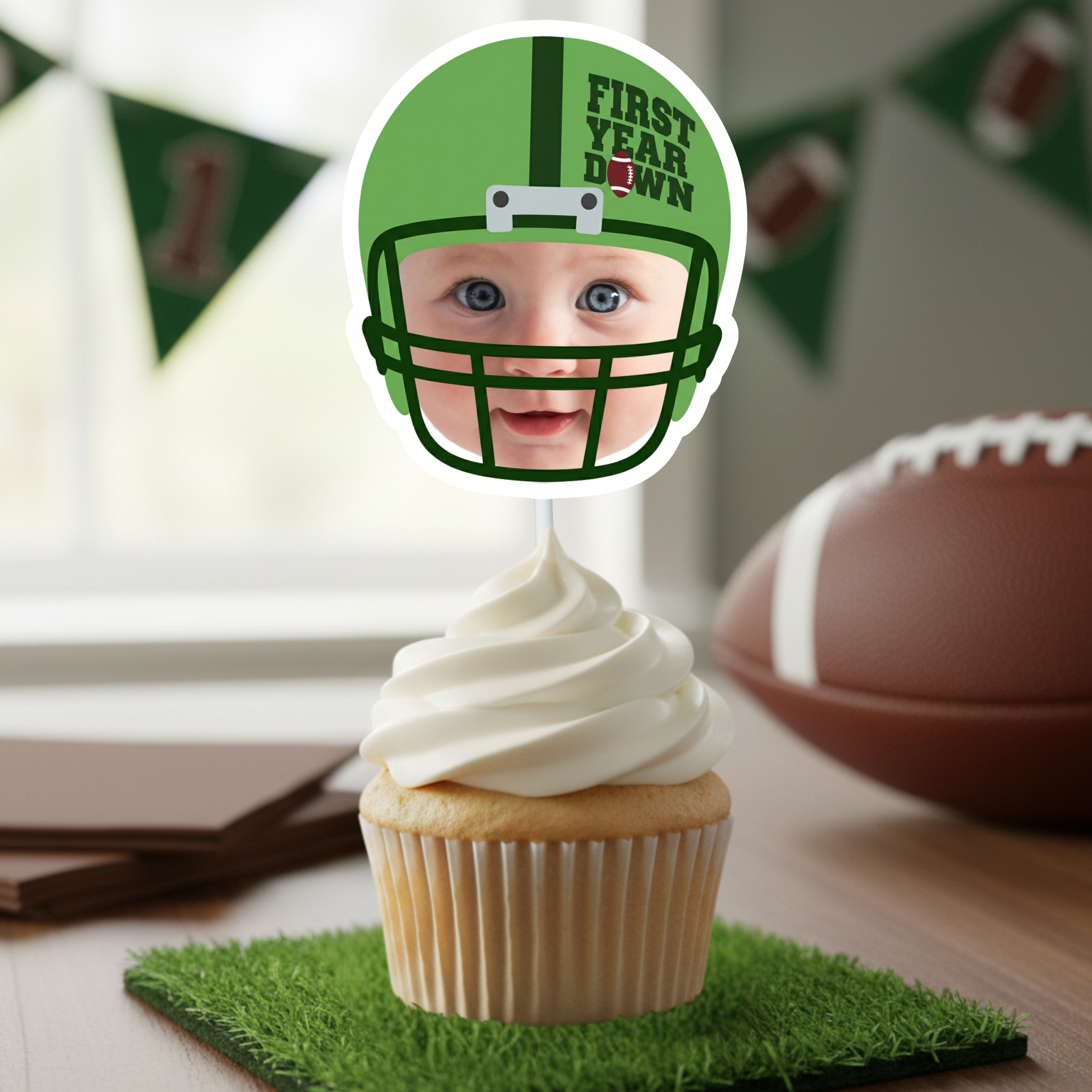 Close up of a green football helmet First Year Down personalized cupcake topper featuring baby face photo design displayed on white frosted cupcake. Football first birthday party decoration styled with football and green banner background for sports themed one year old birthday celebration. Displaying premium quality print resolution. 