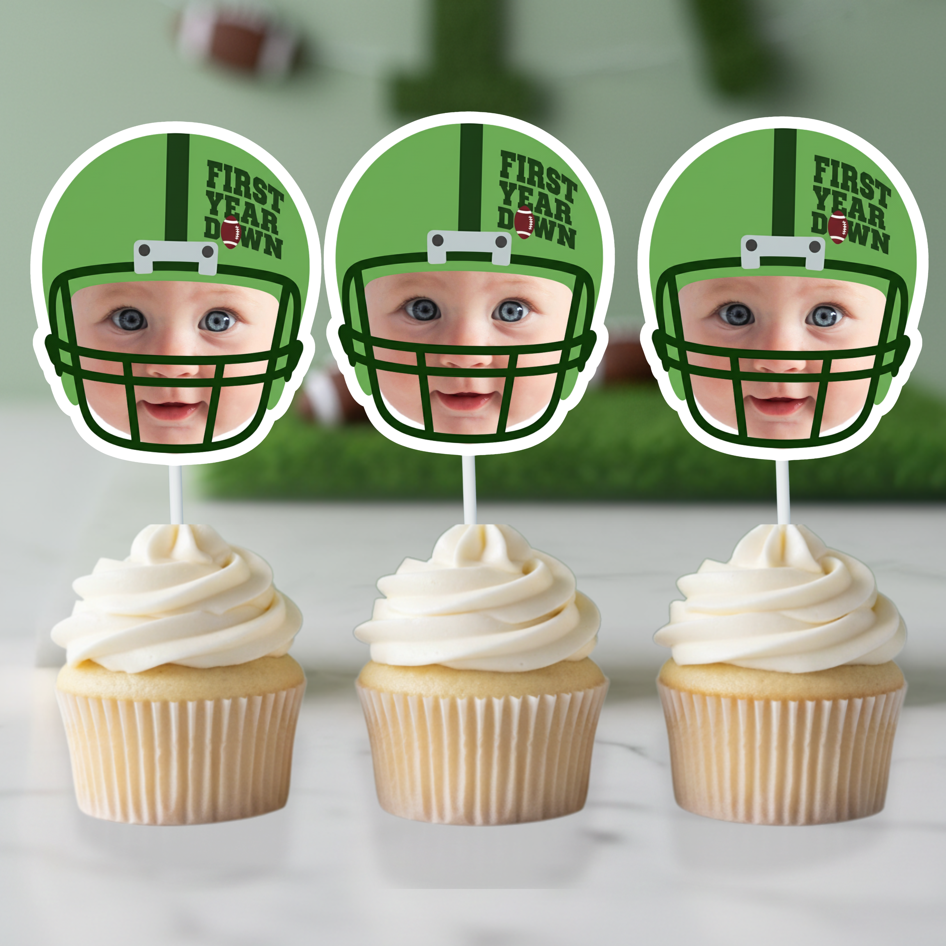 Three green football helmet First Year Down personalized photo cupcake toppers displayed on vanilla cupcakes with smooth white buttercream frosting. Sports themed football 1st birthday party decorations styled for a baby boy dessert table with custom team color helmet design.