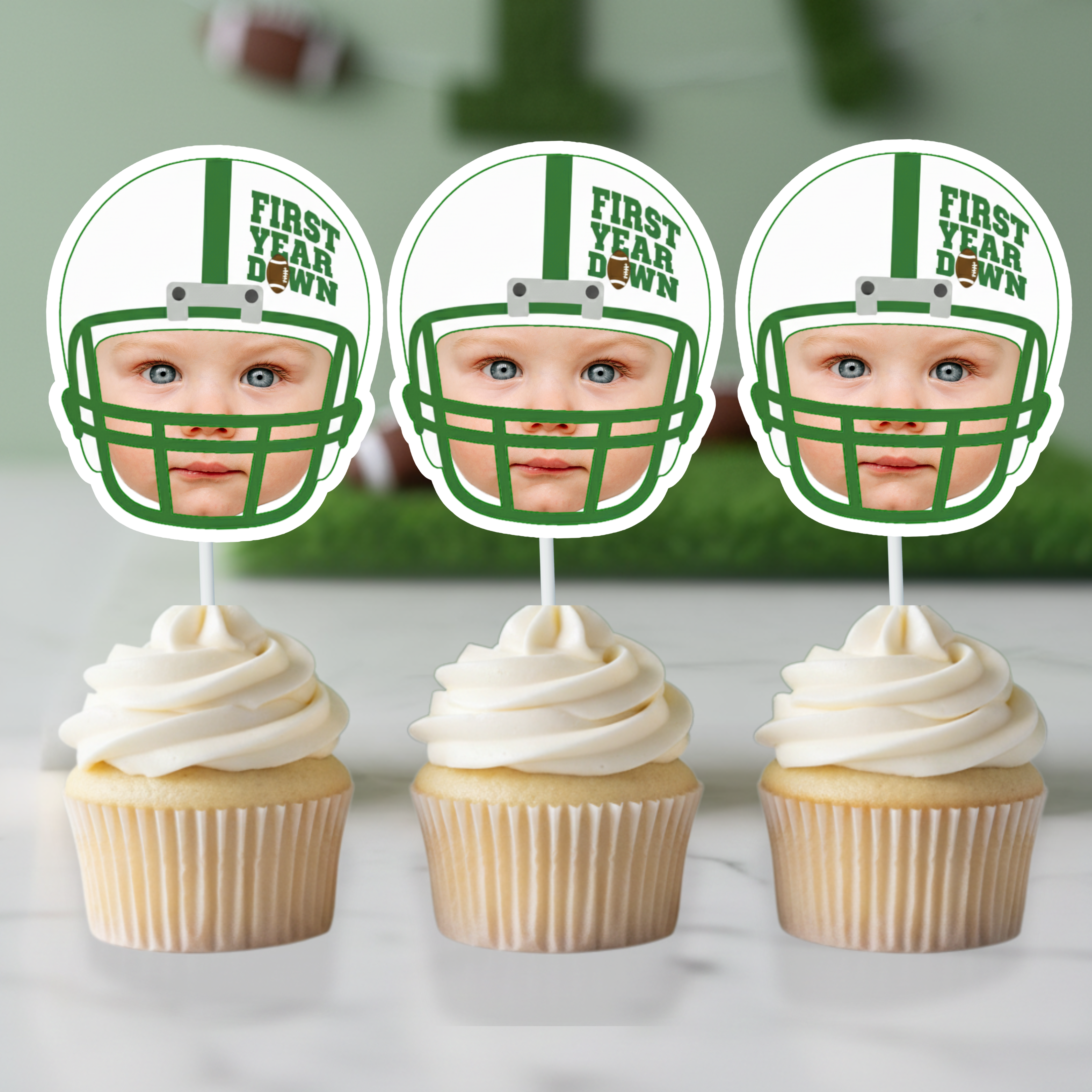 Three green and white football helmet First Year Down personalized photo cupcake toppers displayed on vanilla cupcakes with white buttercream frosting. Styled for football first birthday dessert table with blurred turf background and sports party decor for baby one year celebration.