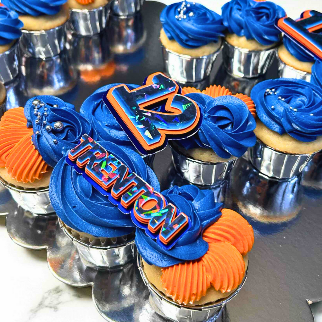 Cupcakes with blue and orange frosting, featuring decorative letters and numbers on a reflective surface.