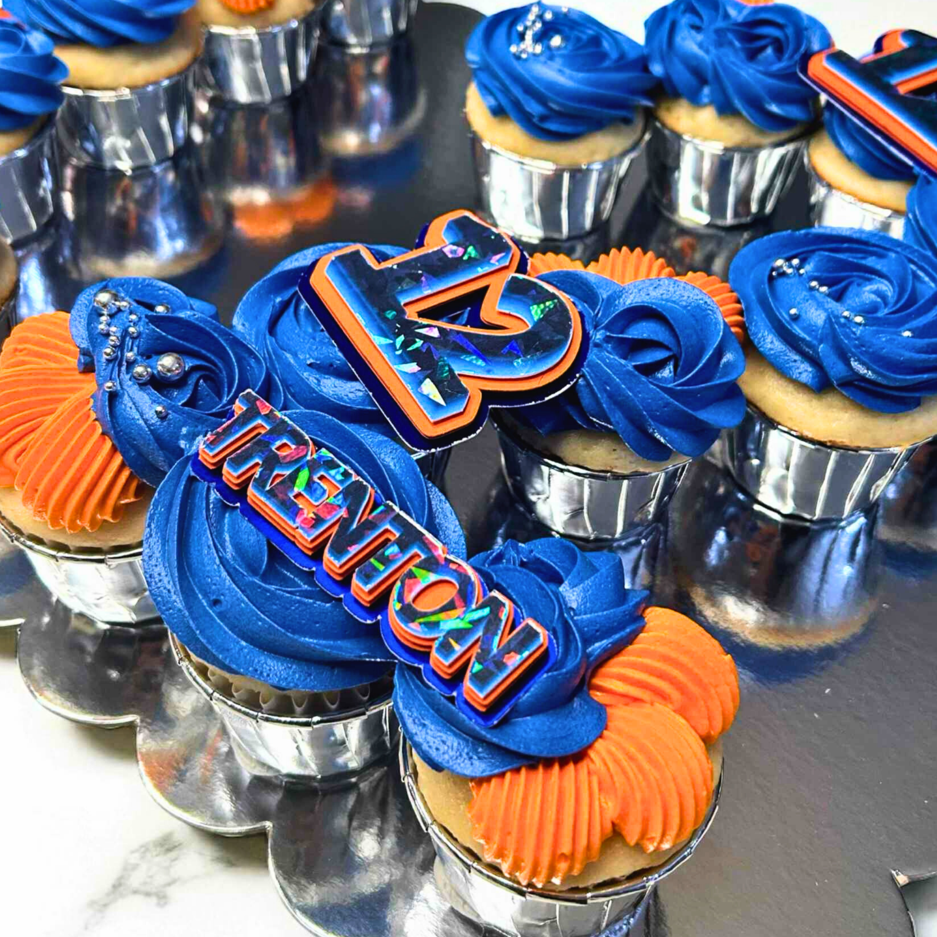 Cupcakes with blue and orange frosting, featuring decorative letters and numbers on a reflective surface.