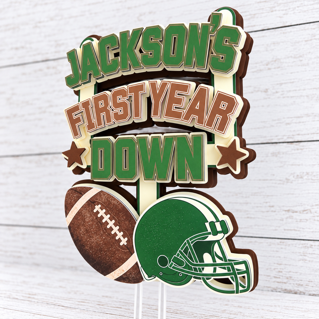 Angled view of handmade layered First Year Down football cake topper showing dimensional cardstock construction in green brown cream and white