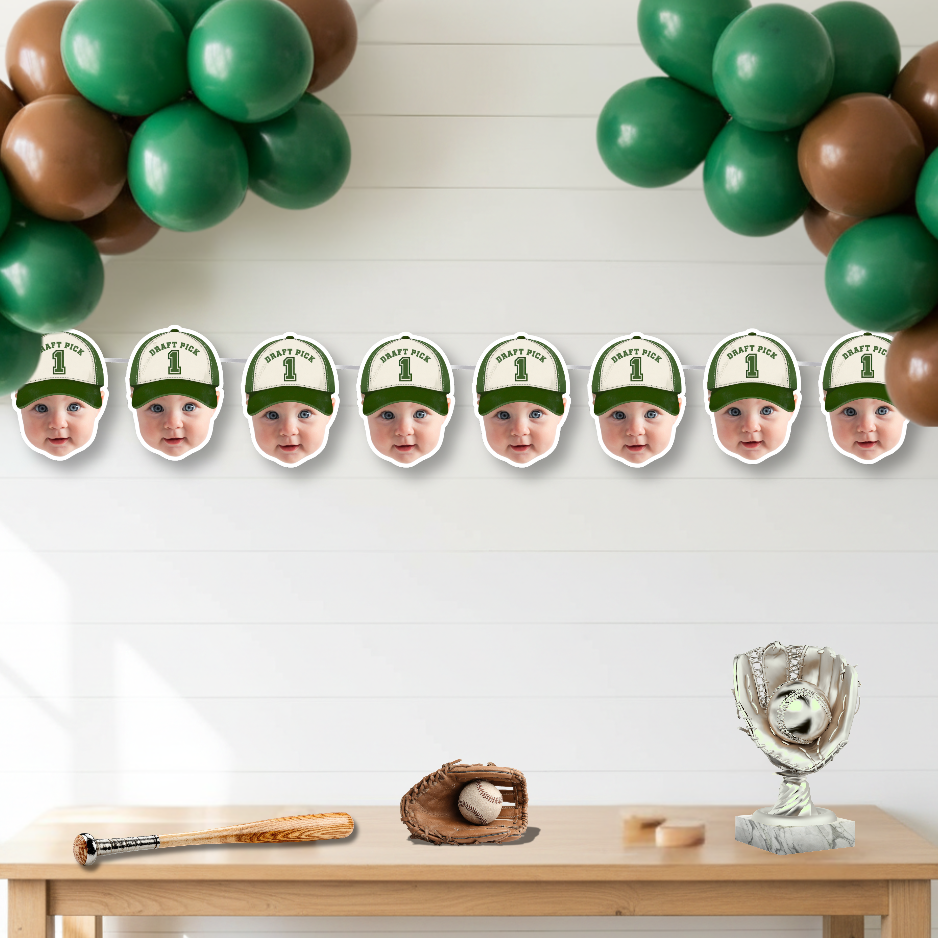 Custom little slugger baseball photo banner featuring Draft Pick 1 Rookie of the Year design displayed beneath green and brown balloon garland, styled as sports birthday backdrop decor for a baseball first birthday celebration.