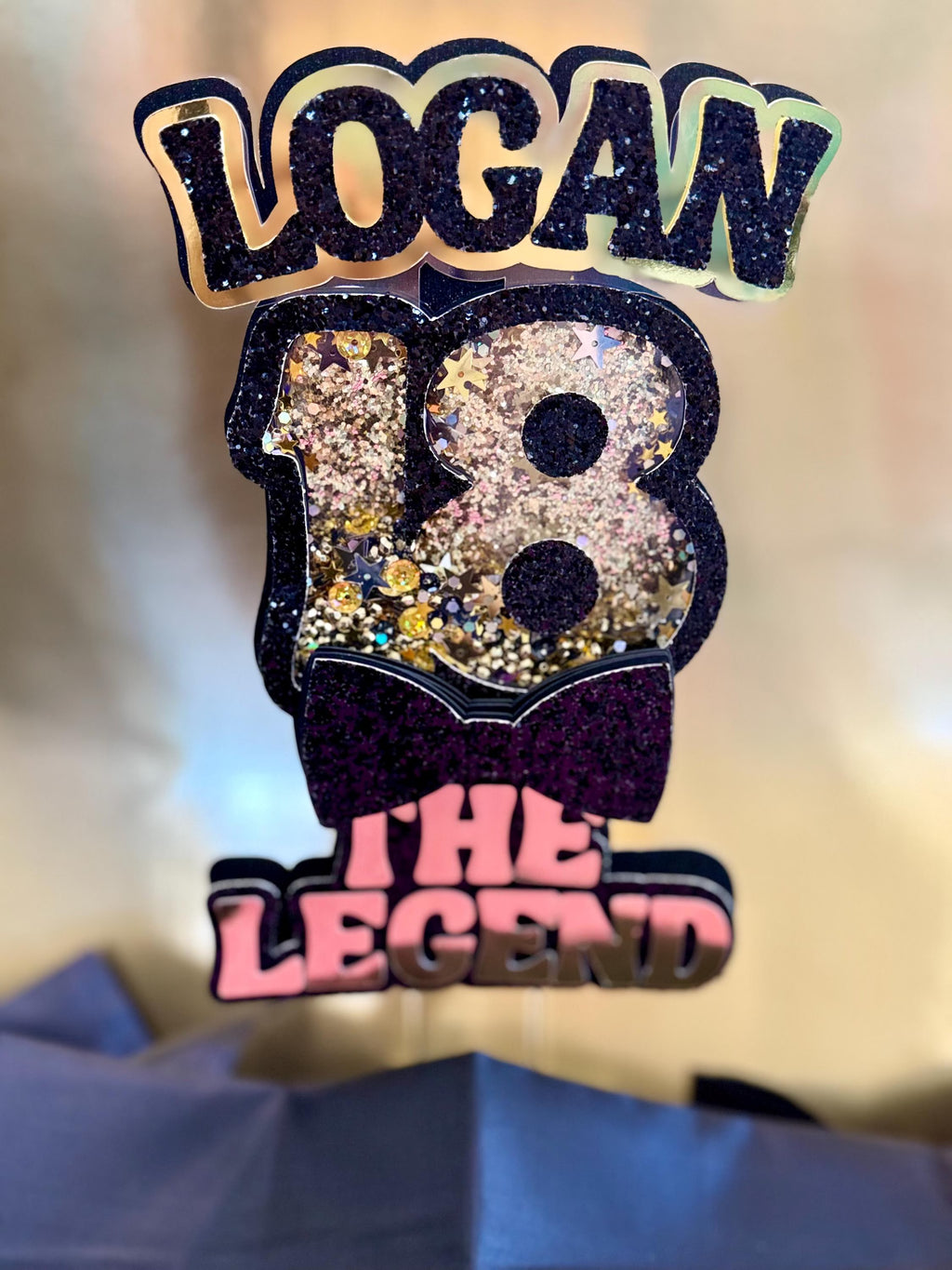 Decorative Gold and Black birthday Cake Topper with 'LOGAN 18 THE LEGEND' text on a blurred background