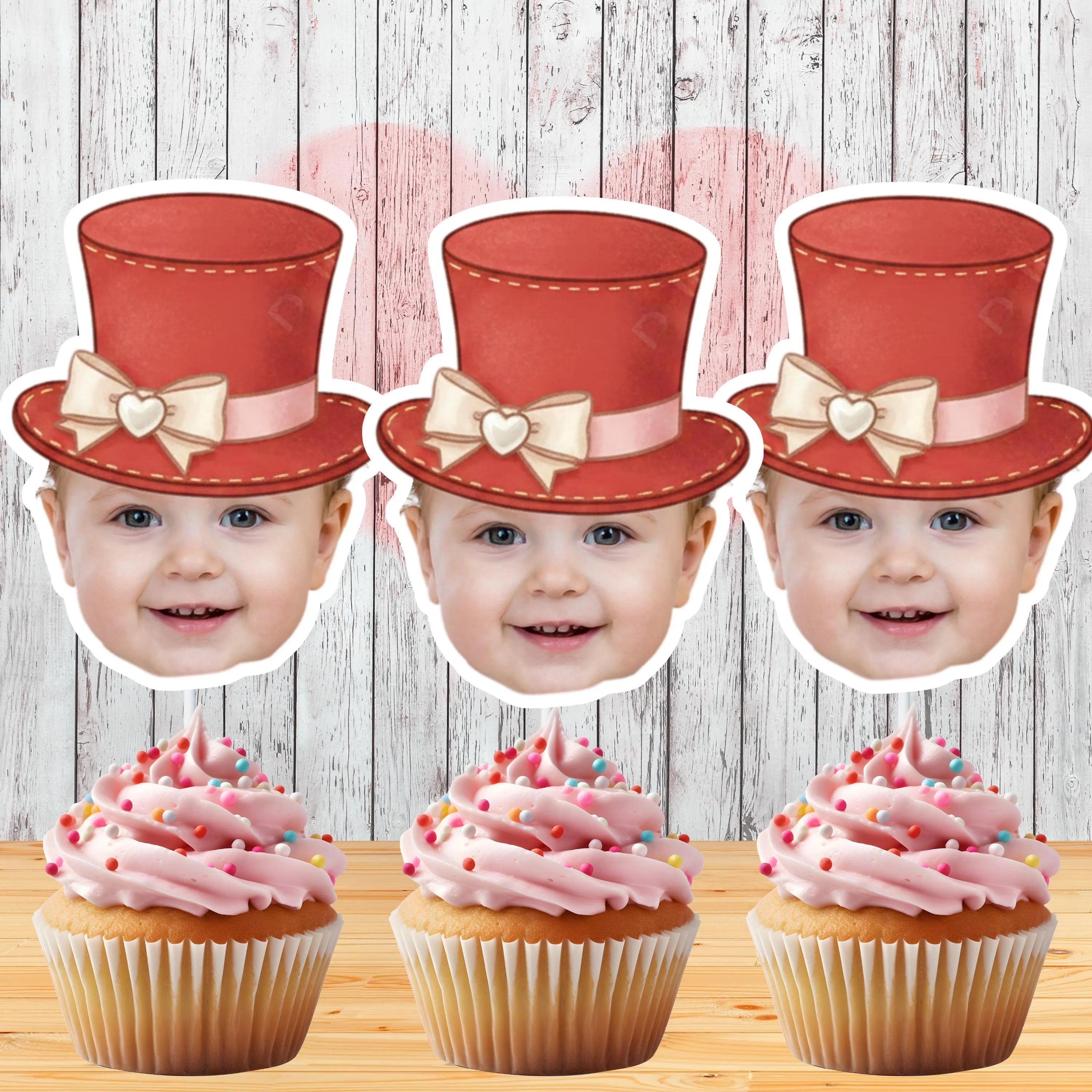 Three children's faces with red top hats and pink ribbons, above cupcakes with pink frosting and sprinkles on a wooden surface.