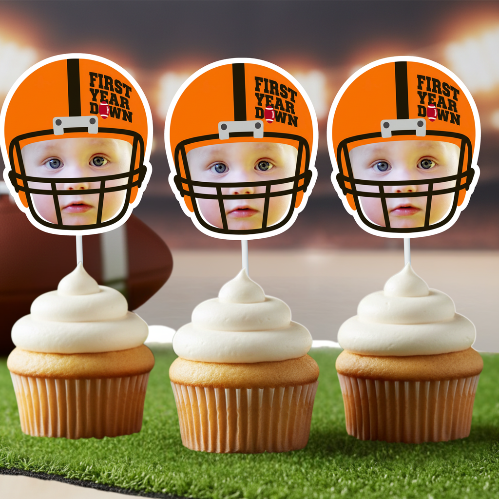 Three orange football helmet First Year Down photo cupcake toppers displayed on vanilla cupcakes with white frosting styled on green turf with stadium lights background. Football first birthday game day party decorations for one year old sports themed celebration.