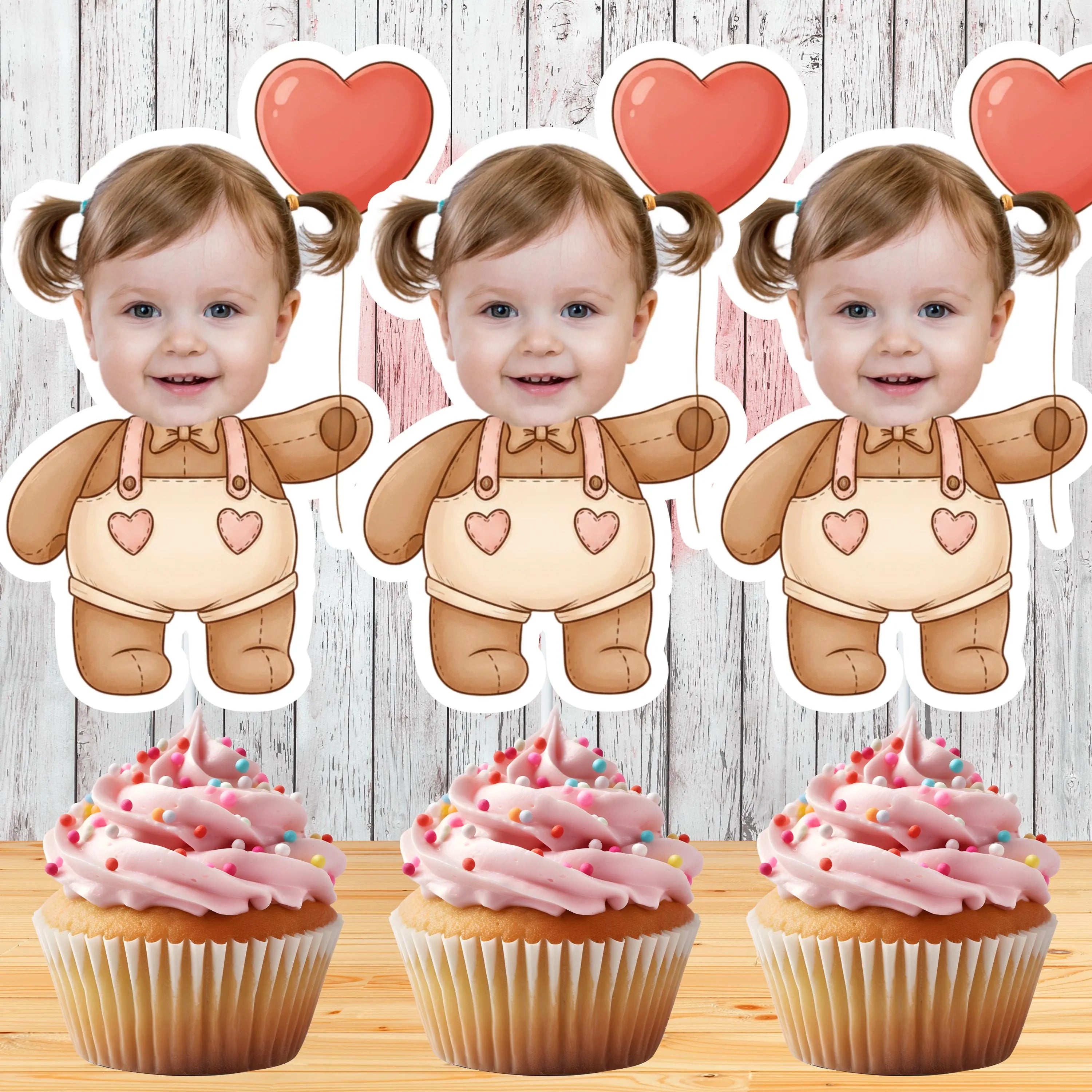 Three identical cupcakes of a baby in a onesie with heart balloons above, next to three cupcakes with pink frosting and sprinkles on a wooden surface.