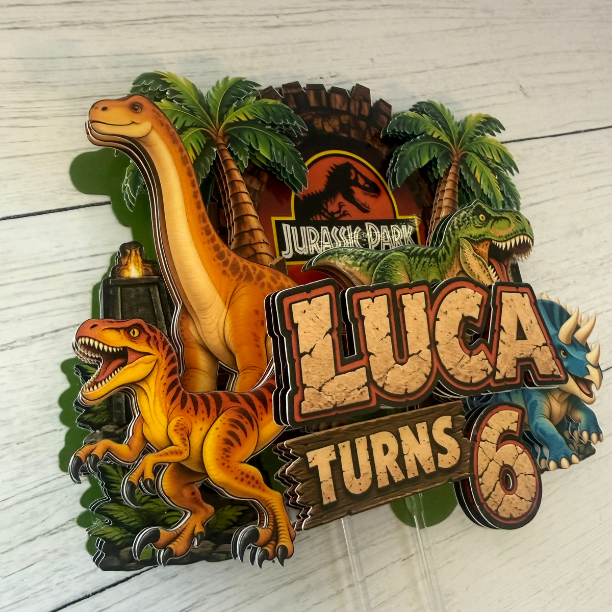 Personalized sixth birthday dinosaur cake topper for Luca featuring Jurassic Park inspired jungle scenery palm trees velociraptor long neck dinosaur triceratops and layered number six details