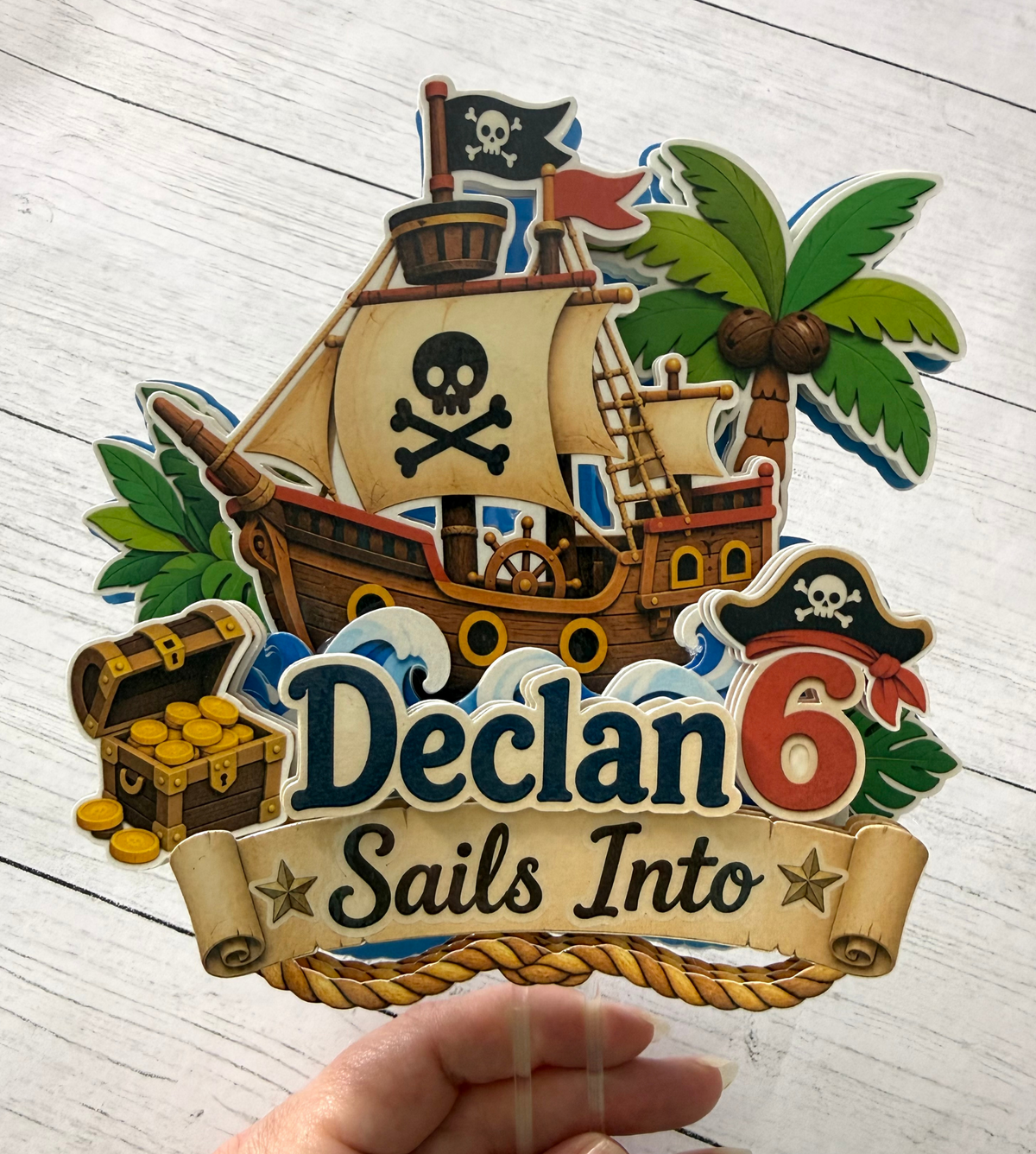 Personalized pirate birthday cake topper held by hand to show approximate scale and layered cardstock details with custom name and age.