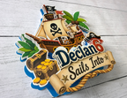 Close up angled view of personalized pirate birthday cake topper showing raised layered cardstock construction pirate ship treasure chest and custom name and age.