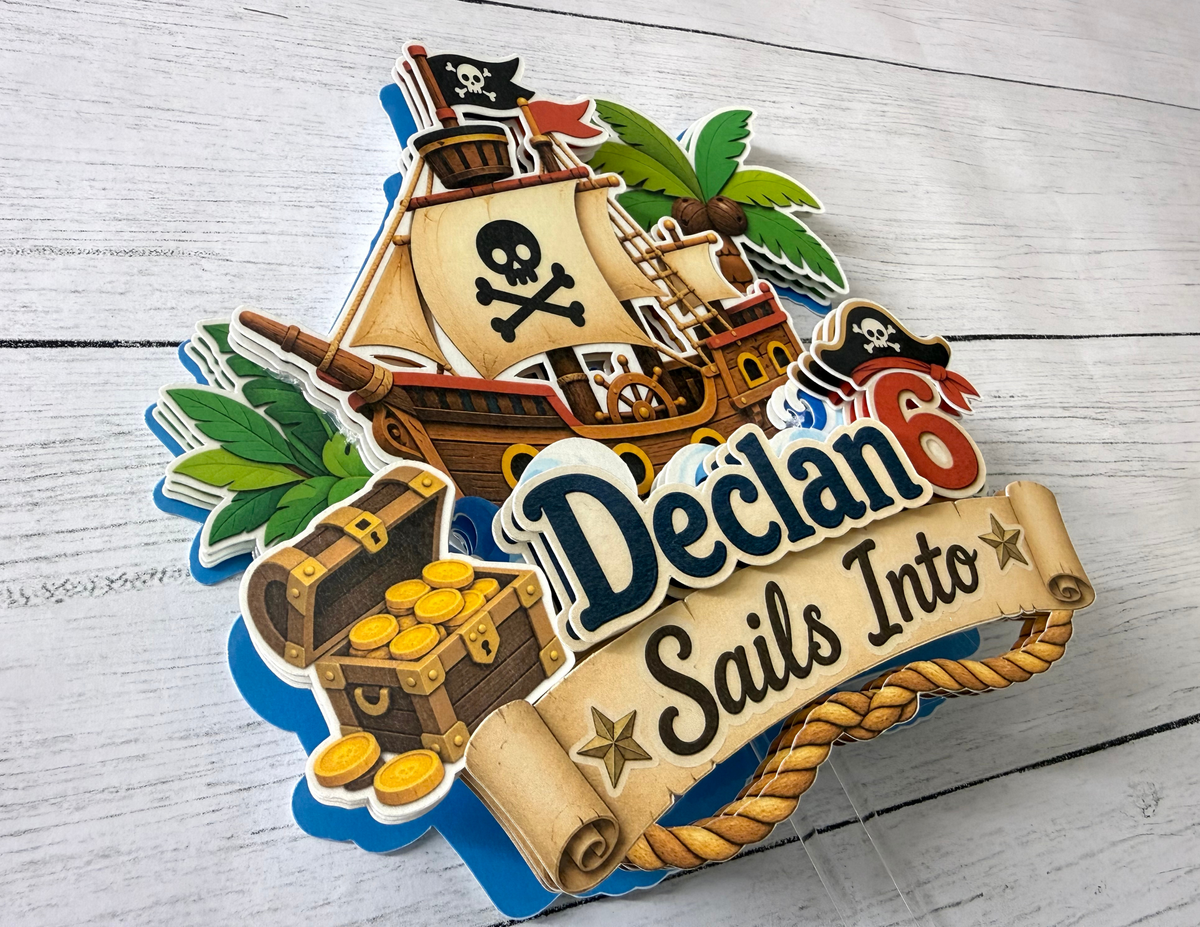 Close up angled view of personalized pirate birthday cake topper showing raised layered cardstock construction pirate ship treasure chest and custom name and age.