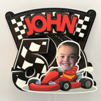 Closeup of personalized racing birthday cake topper with child photo John name age five red go kart checkered flags and black glitter background