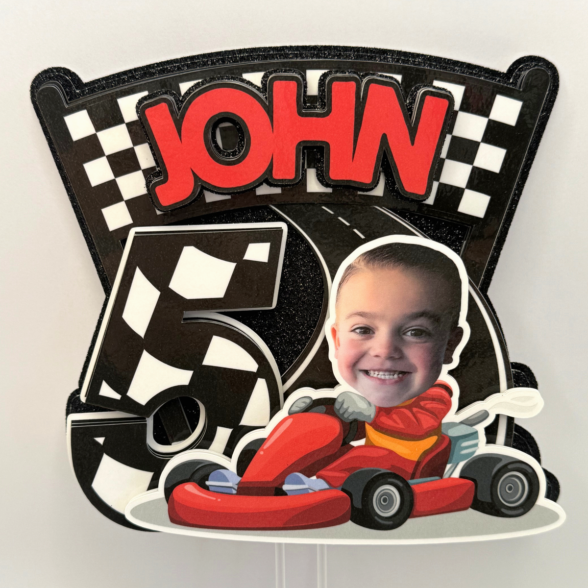 Closeup of personalized racing birthday cake topper with child photo John name age five red go kart checkered flags and black glitter background