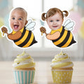 Two cupcake toppers with children's faces and bee designs on a blurred background.