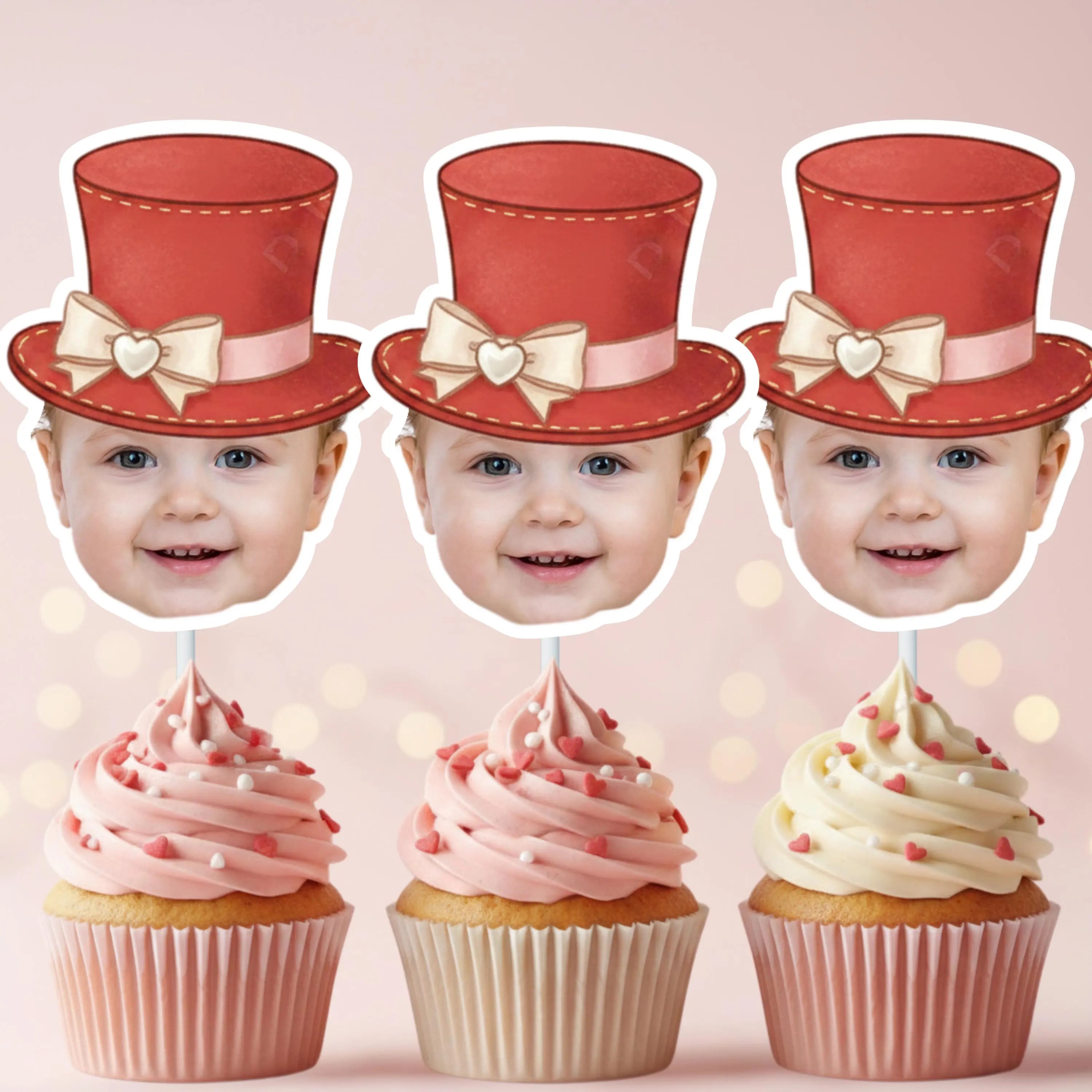 Three cupcakes with pink frosting and white ribbons, each topped with a baby's face wearing a red top hat with a bow.