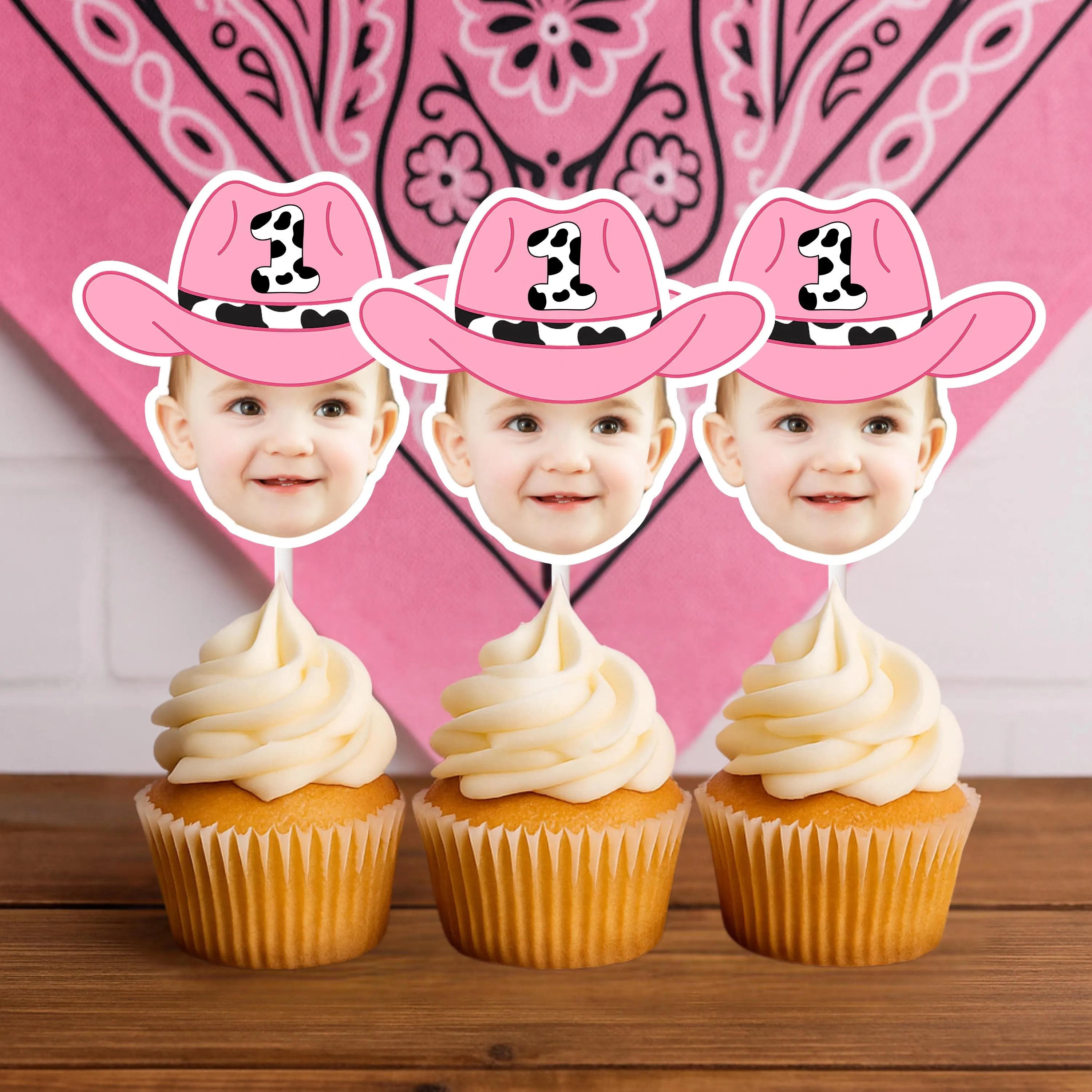Three cupcakes with pink cowboy hats and faces on top against a pink bandana patterned background