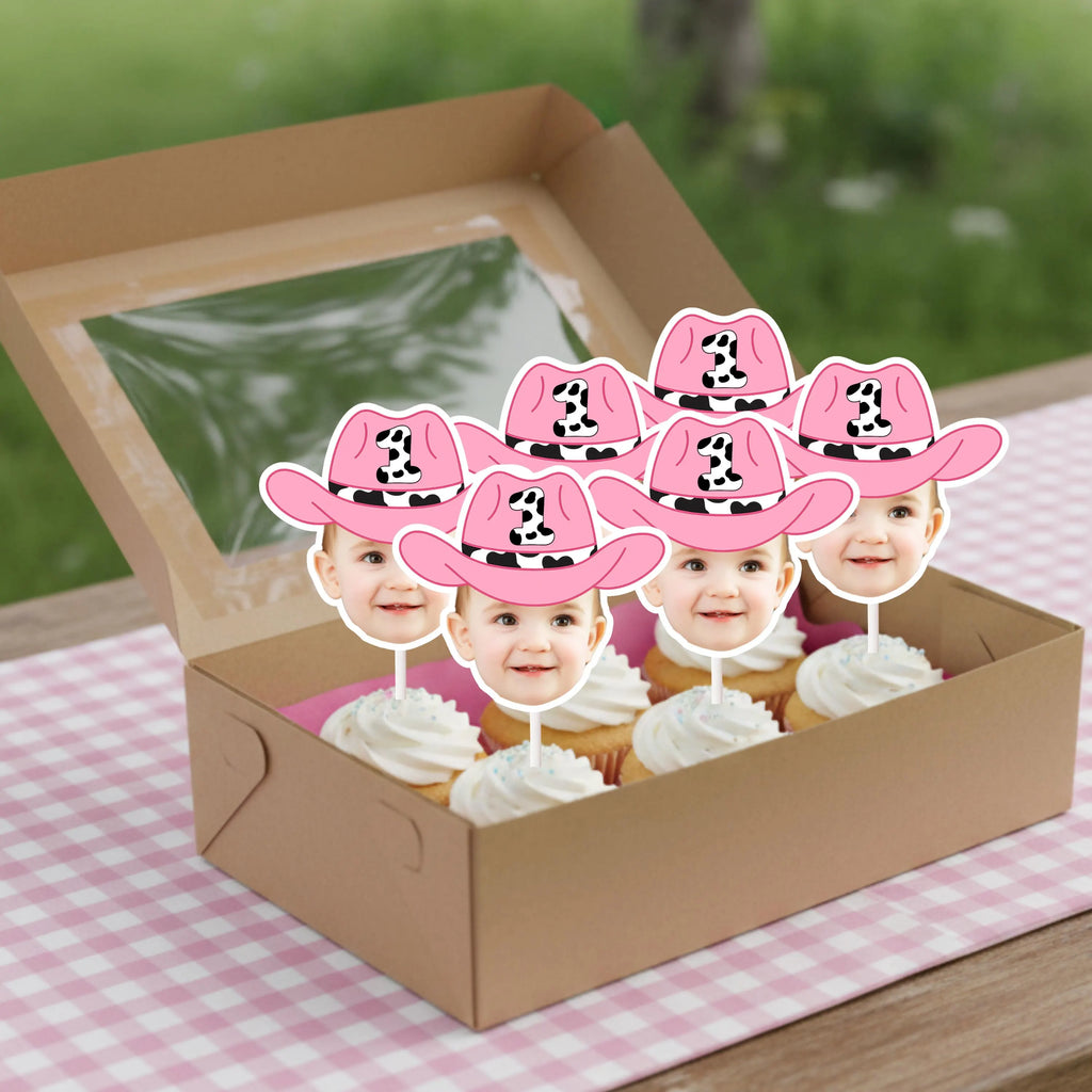 Cupcakes with pink cowboy hats and face cutouts in a box on a checkered tablecloth.