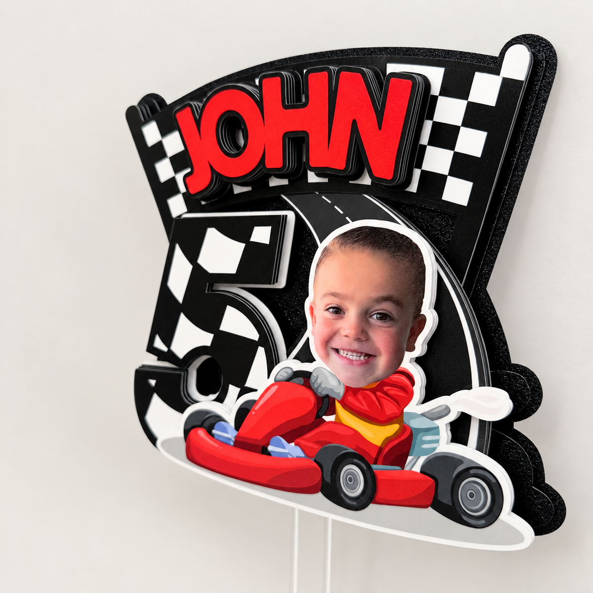 Angled view of layered personalized race car birthday cake topper with child photo name and age in red black white checkered racing colors
