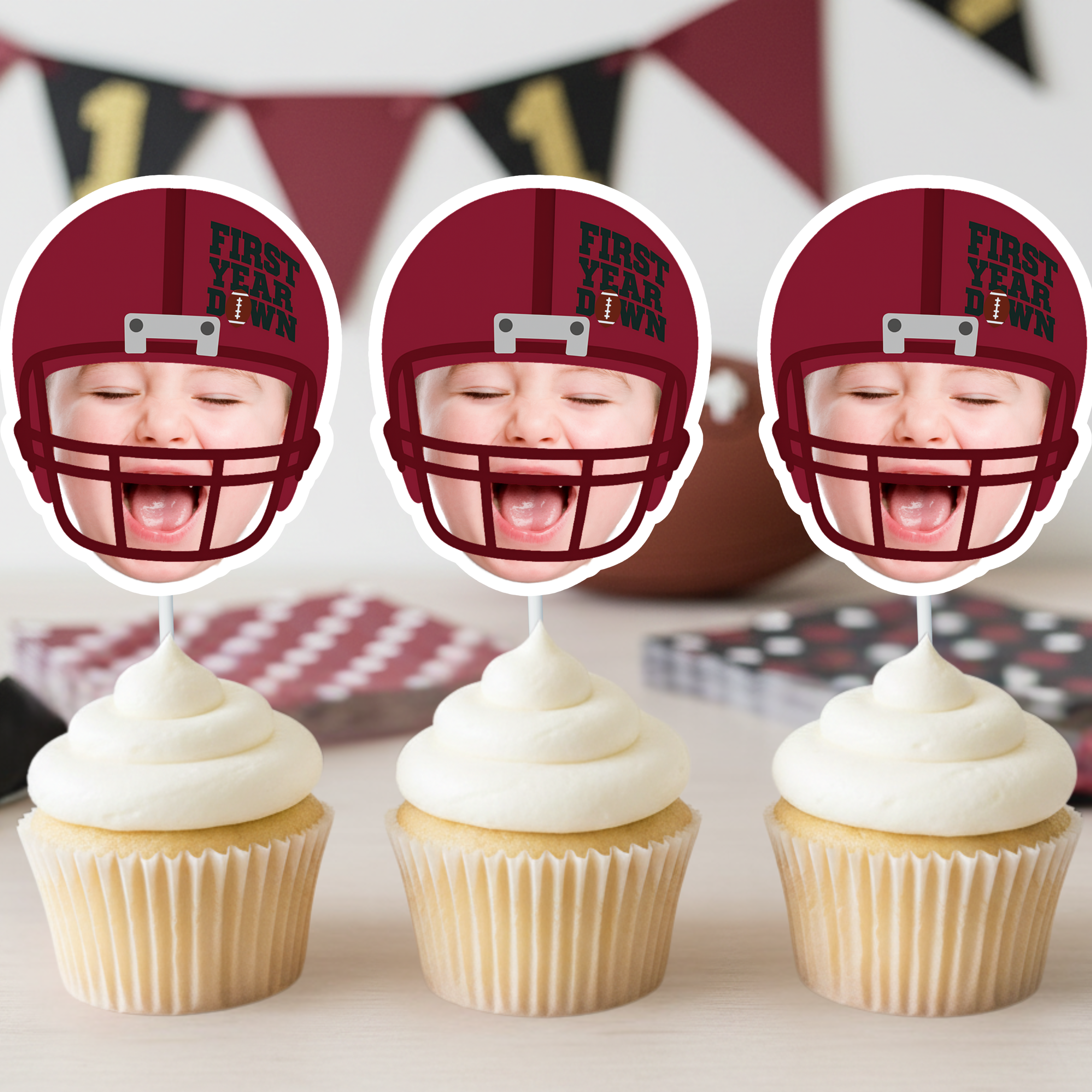 Three red football helmet First Year Down personalized cupcake toppers featuring baby photo design displayed on frosted cupcakes for football themed first birthday party. Burgundy and black team color sports birthday decorations styled with football banner background.