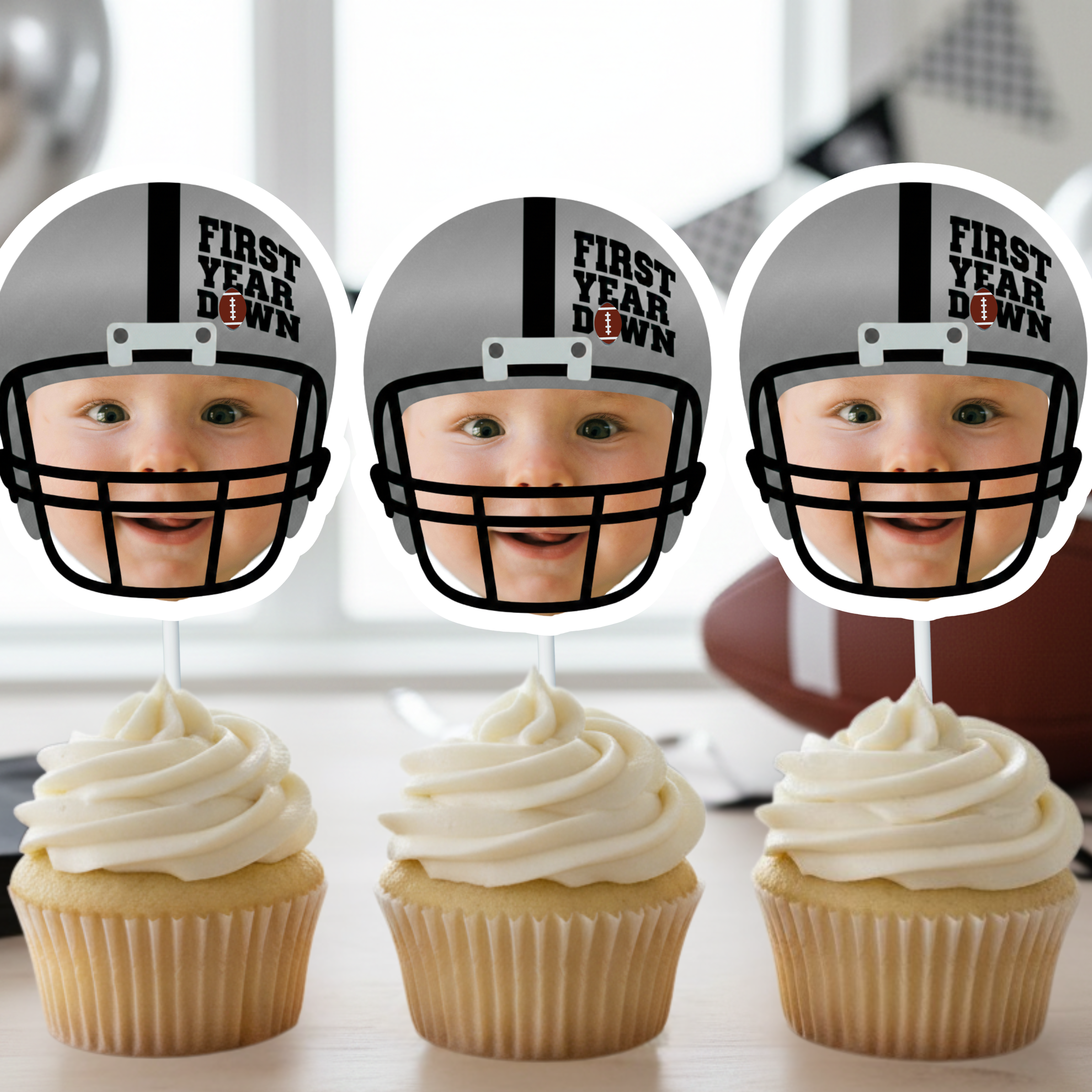 Three silver football helmet First Year Down personalized photo cupcake toppers displayed on vanilla cupcakes with white buttercream frosting. Modern football first birthday party decorations styled with neutral black and white banner and sports themed table setup for baby boy one year old celebration.