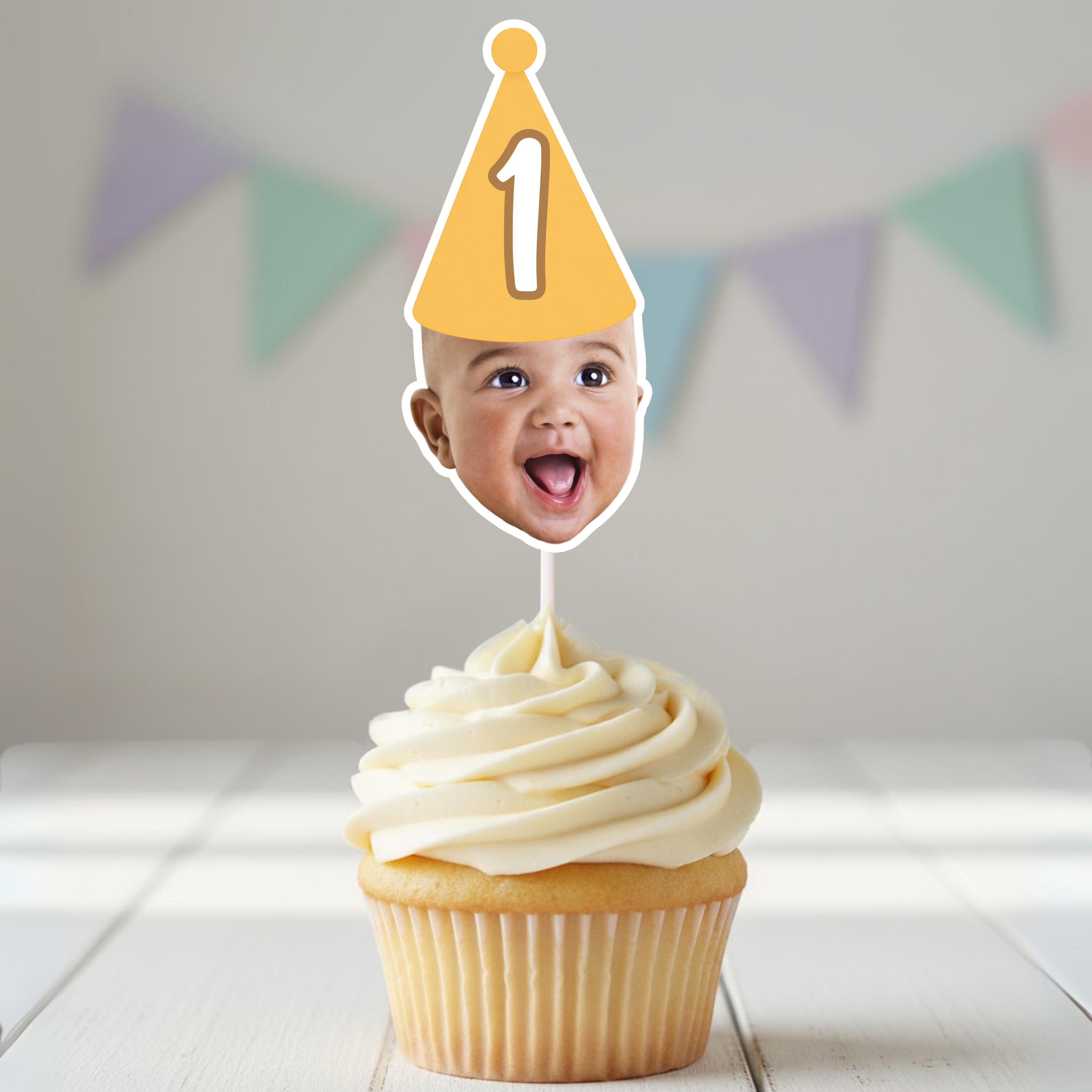 Cupcake with a photo of a baby on top, wearing a party hat with the number 1, against a light background with colorful flags.
