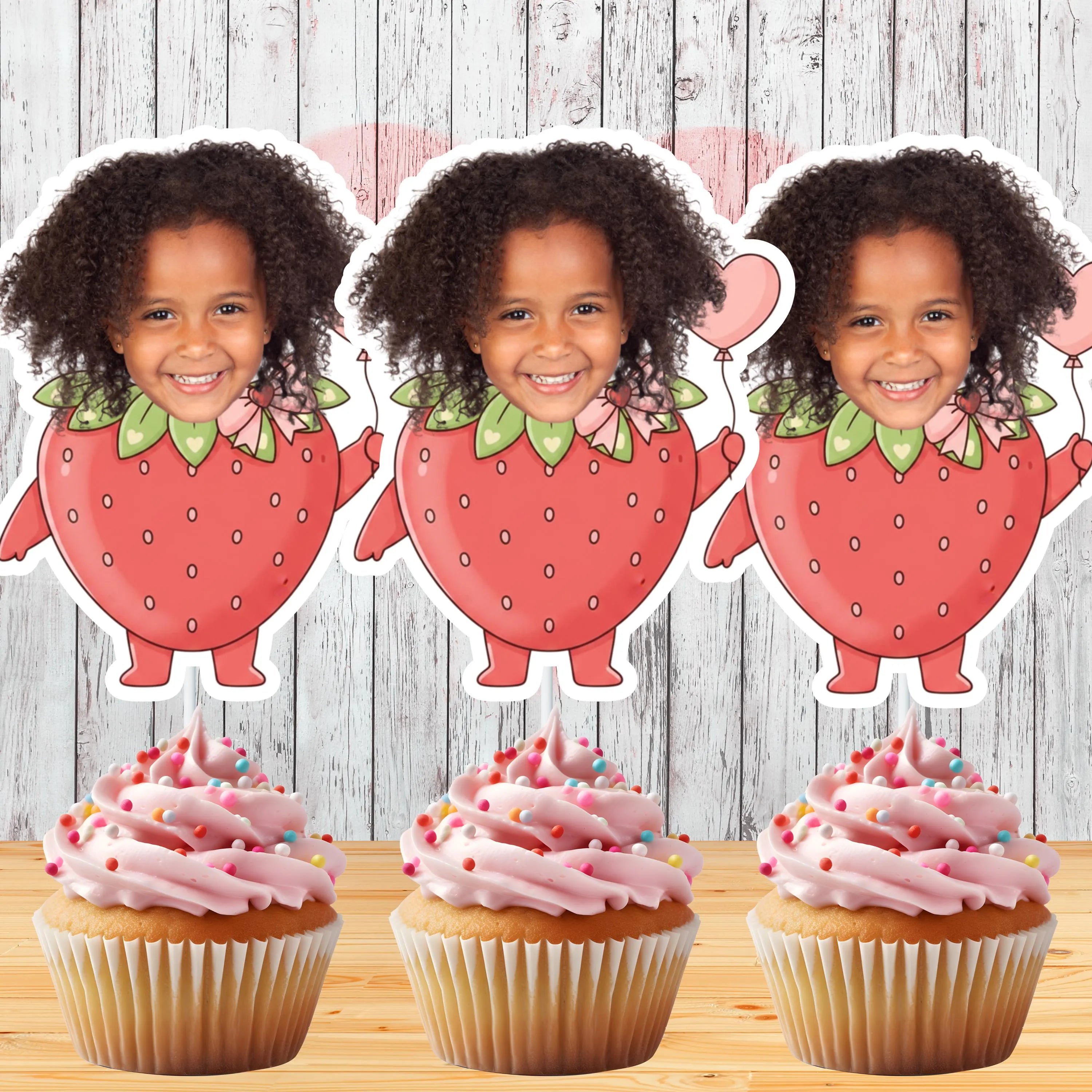 Three strawberry-shaped cutouts with a child's face on top, placed above cupcakes with pink frosting and sprinkles.