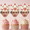 Three cupcakes with pink frosting and heart decorations, each topped with a cardboard crown featuring hearts.