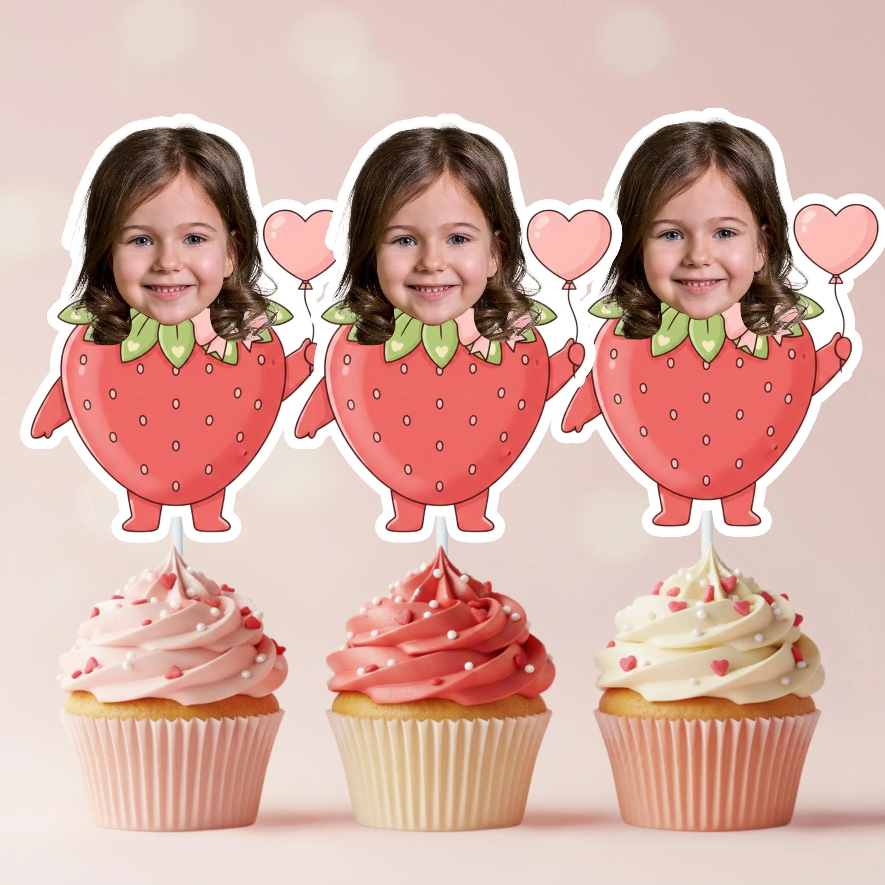 Three strawberry-shaped cupcake toppers with a child's face and pink balloons, placed on cupcakes with pink frosting.