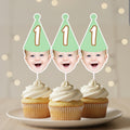 Three cupcakes with cream frosting on a plate, each topped with a baby's face cutout wearing a green party hat with the number '1'.