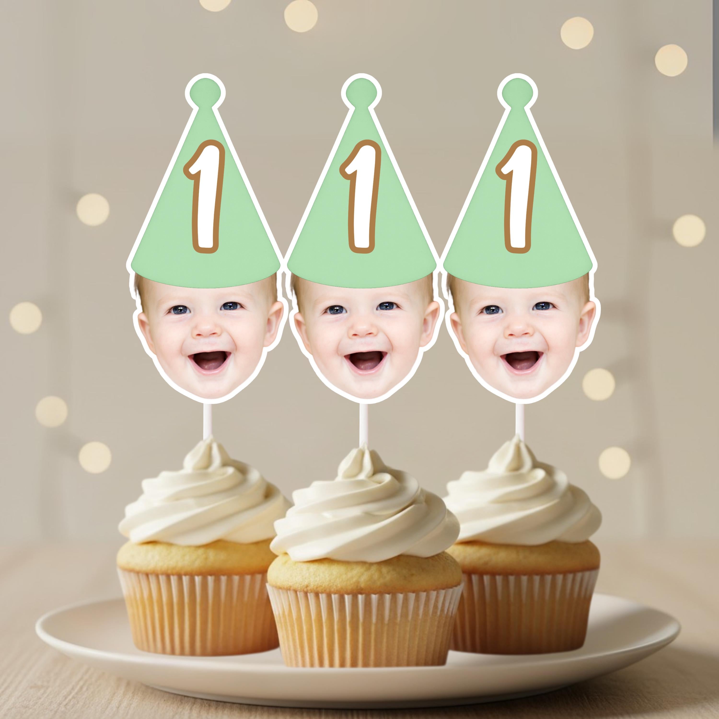 Three cupcakes with cream frosting on a plate, each topped with a baby's face cutout wearing a green party hat with the number '1'.